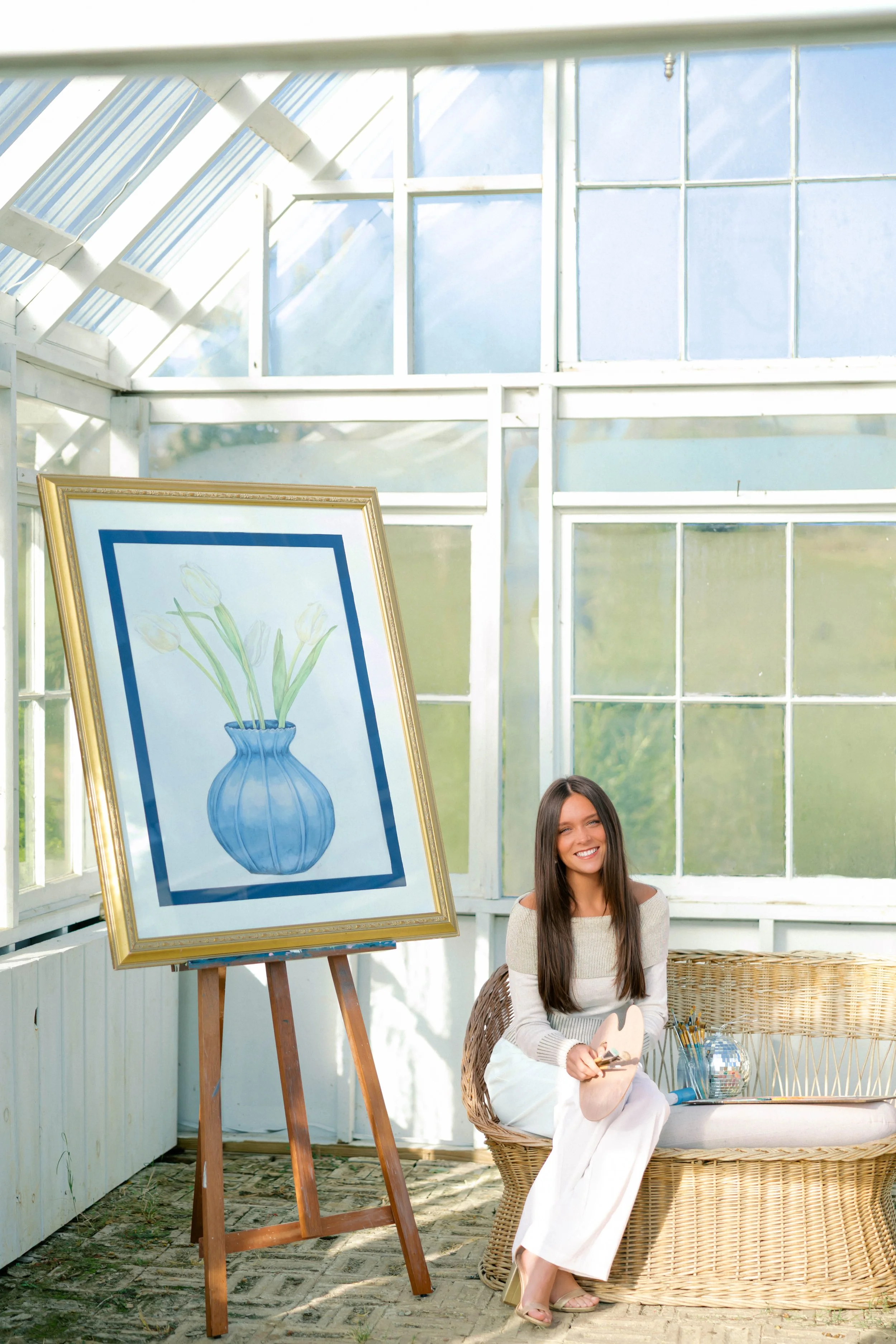 A smiling young woman with long dark hair sitting in a wicker chair in a greenhouse-like space with large windows, next to an easel displaying a watercolor painting of a blue vase with white tulips. She holds a painting palette and brushes, with art supplies on a small table beside her.