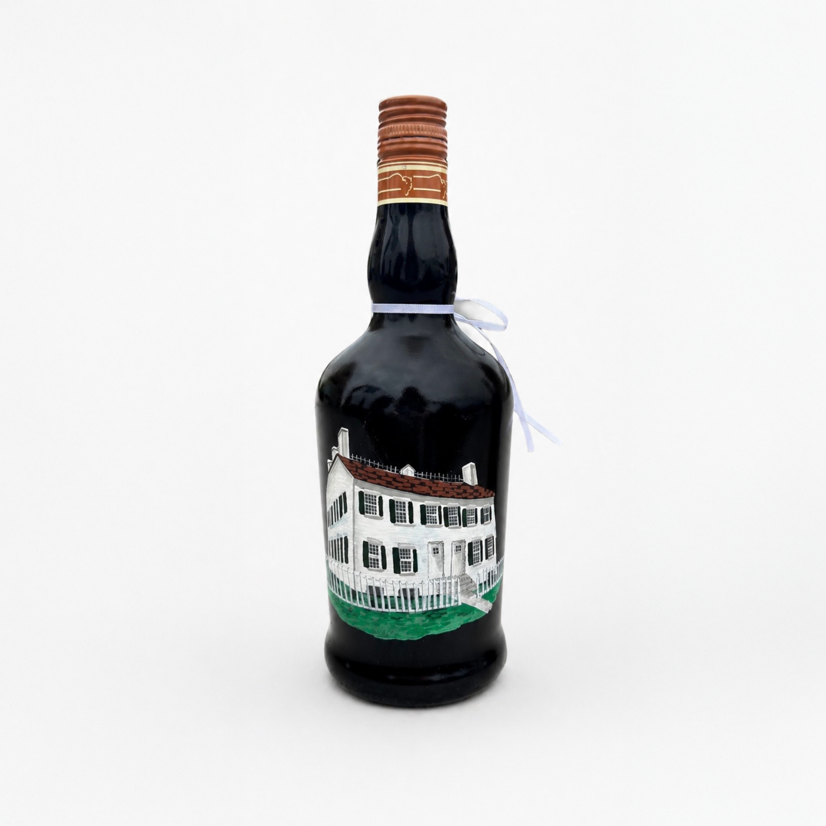 A dark glass bottle with a white ribbon tied around its neck, featuring a label with an illustration of a white building with a red roof, black shutters, surrounded by greenery.