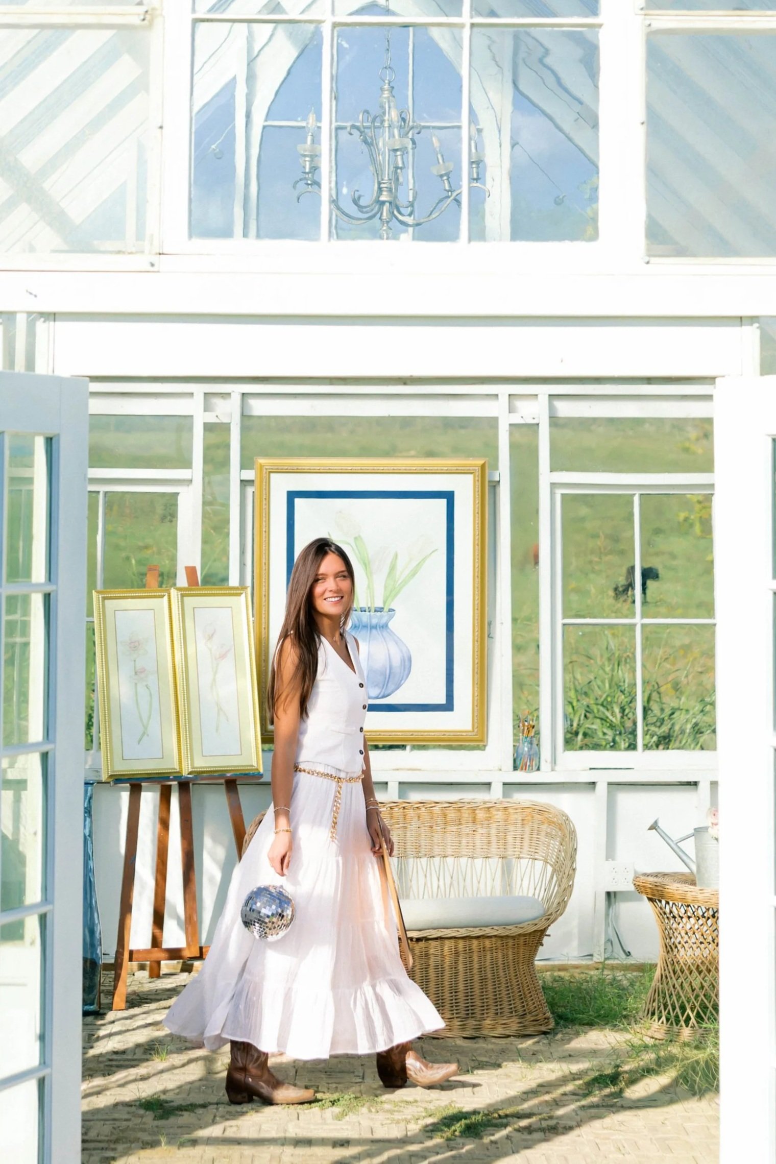 A woman in a white dress and brown cowboy boots standing in front of a garden scene inside a glass greenhouse, with artwork, wicker furniture, and a disco ball nearby.
