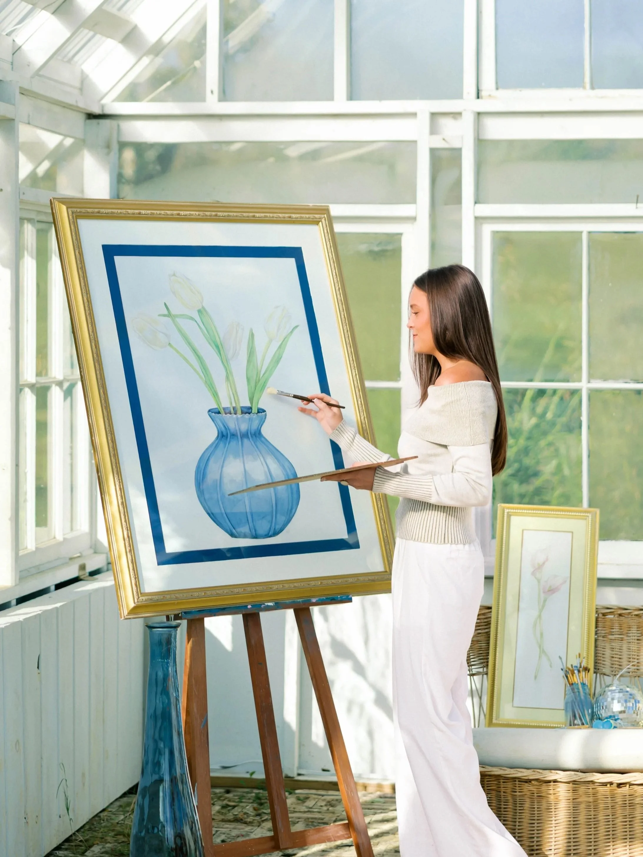 A young woman painting a still life of tulips in a blue vase on a large canvas inside a bright, airy greenhouse with large windows.