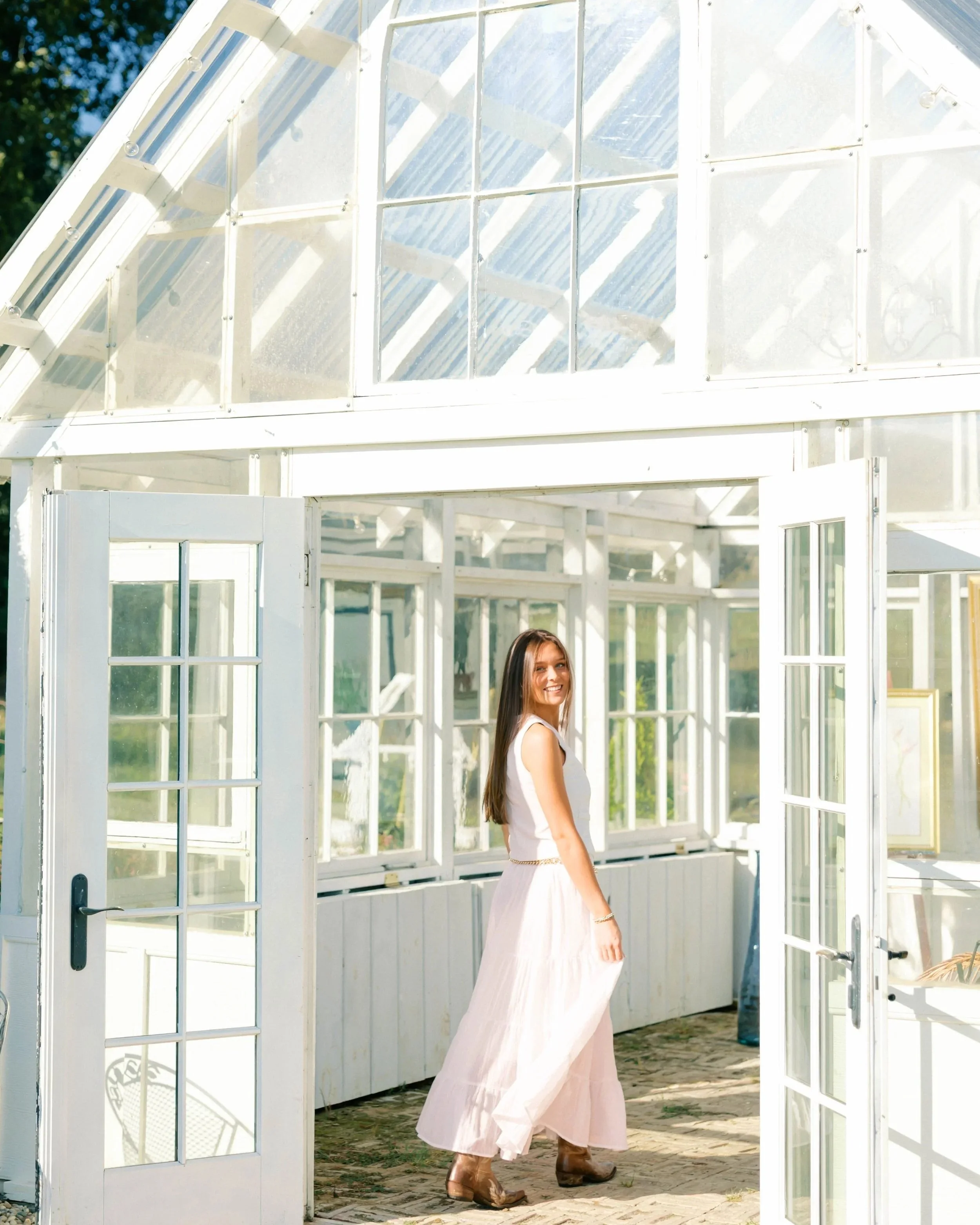 A woman with long brown hair and a white dress with a flowing skirt, wearing cowboy boots, standing in a bright greenhouse with glass windows and doors, smiling and looking towards the camera.