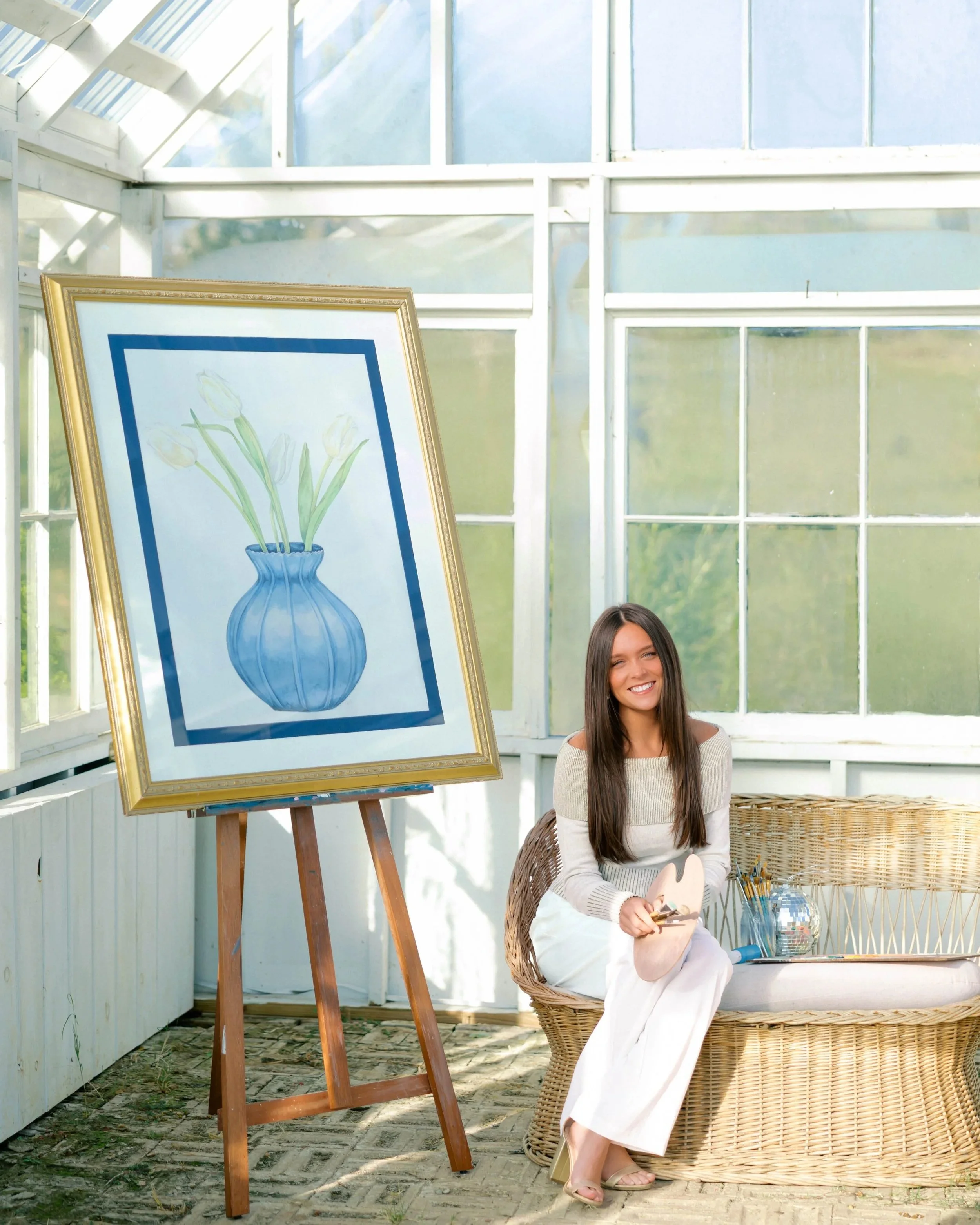 A woman with long dark hair and a beige sweater sits on a wicker chair inside a bright greenhouse, holding a small artist's palette. Behind her, a framed watercolor painting of white tulips in a blue vase is displayed on an easel. Sunlight filters through the glass panels of the greenhouse, casting natural light inside.