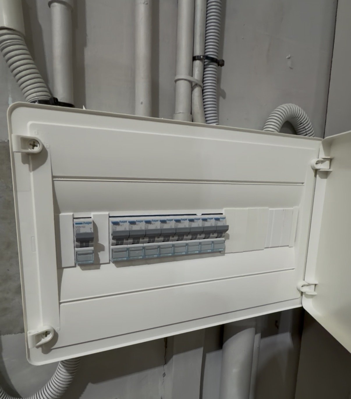 An electrical panel box with multiple circuit breakers inside, mounted on a wall with visible electrical conduits above.