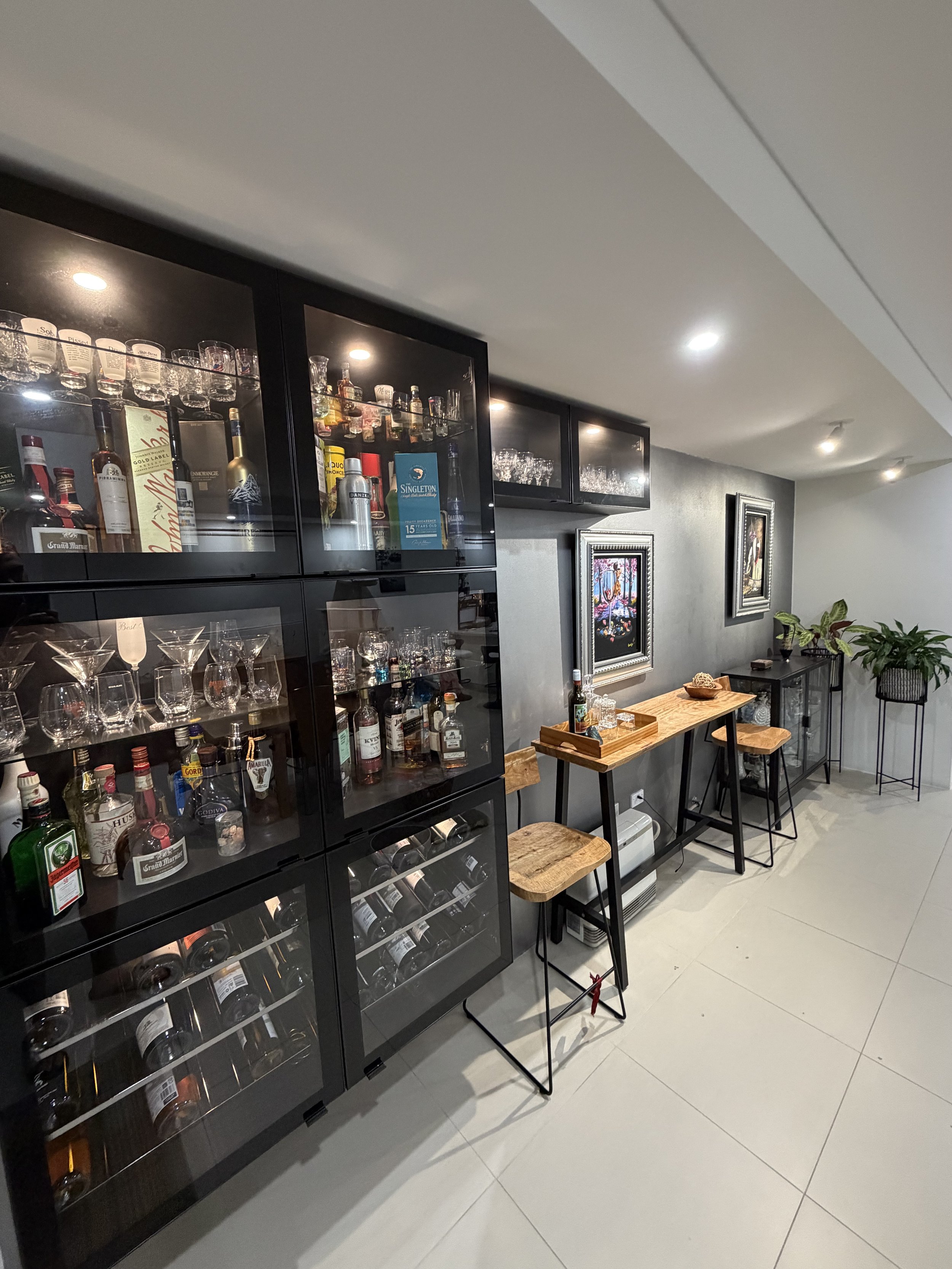 A home bar with glass shelving containing various bottles of alcohol and glassware, accented with artwork and plants, and a small wooden table with bar tools and stools.