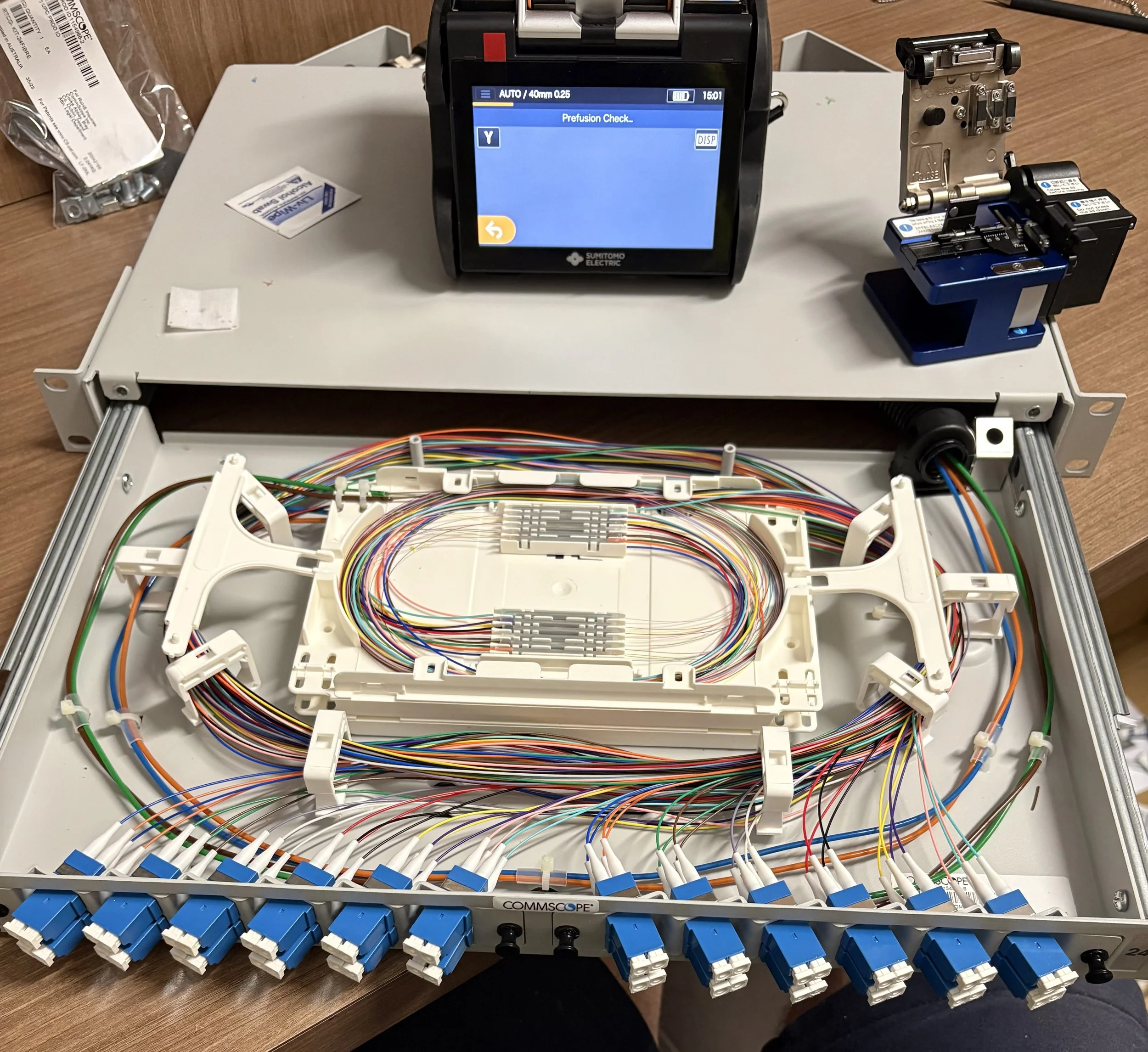 A telecommunications patch panel with numerous colored cables connected to it, a testing device displaying a screen with the message 'Prefusion Check,' and various small parts and tools on a wooden surface.