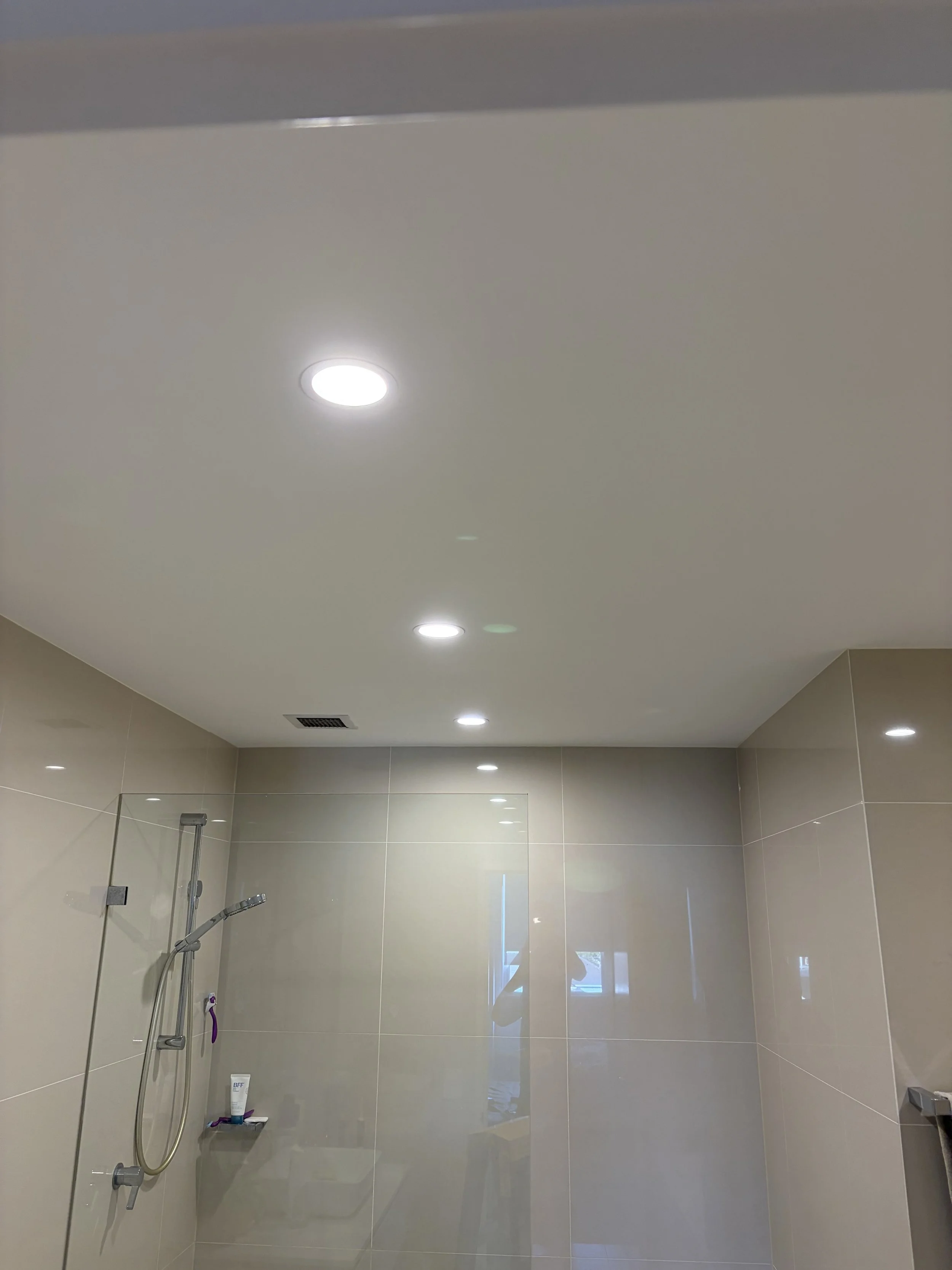 A bathroom shower area with beige tiled walls, a showerhead, a purple and white razor, and a small shelf holding lotion and soap. Recessed ceiling lights illuminate the space.