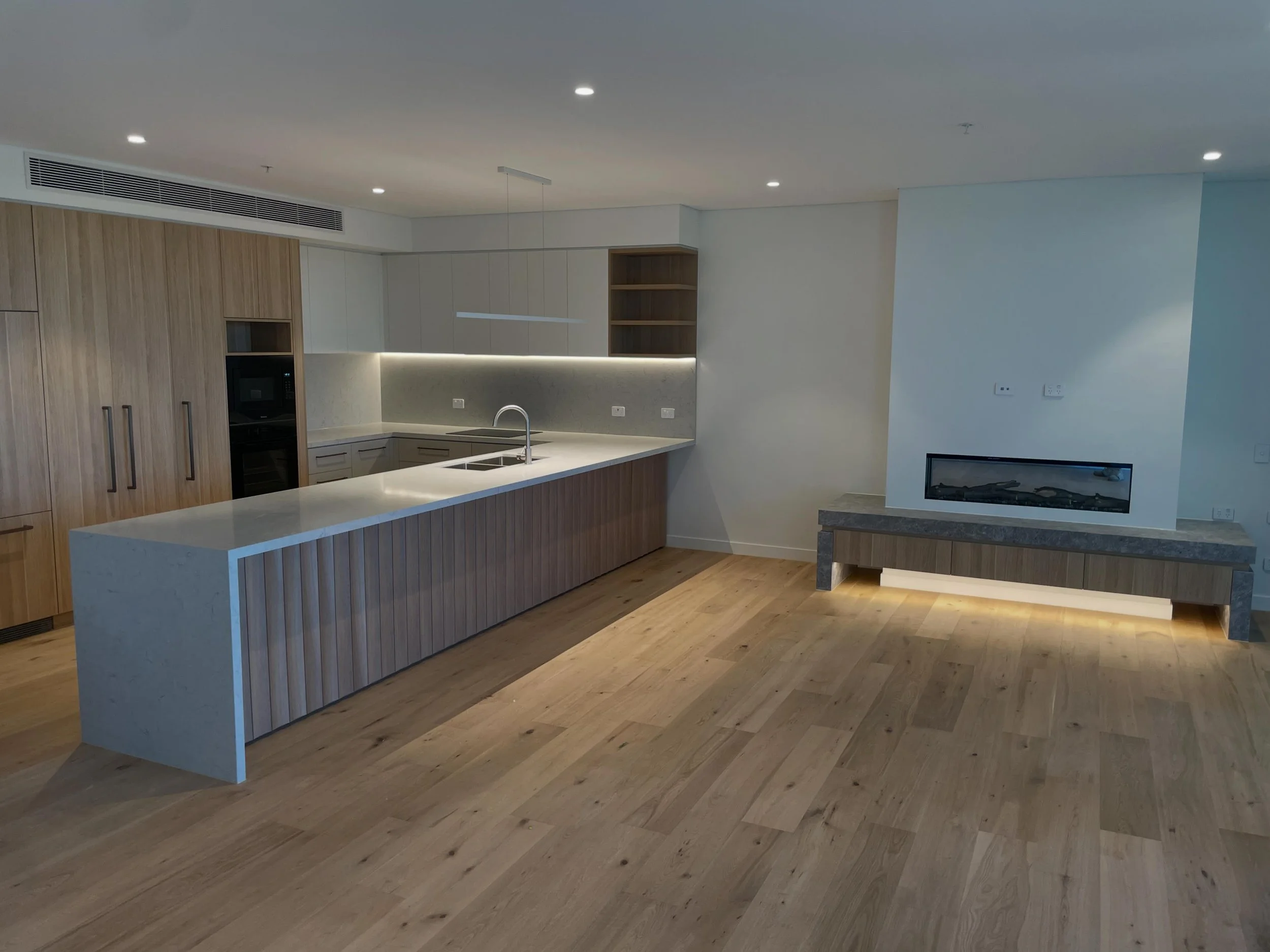 Modern kitchen with light wood cabinets, a white island counter, and a fireplace in the living area.