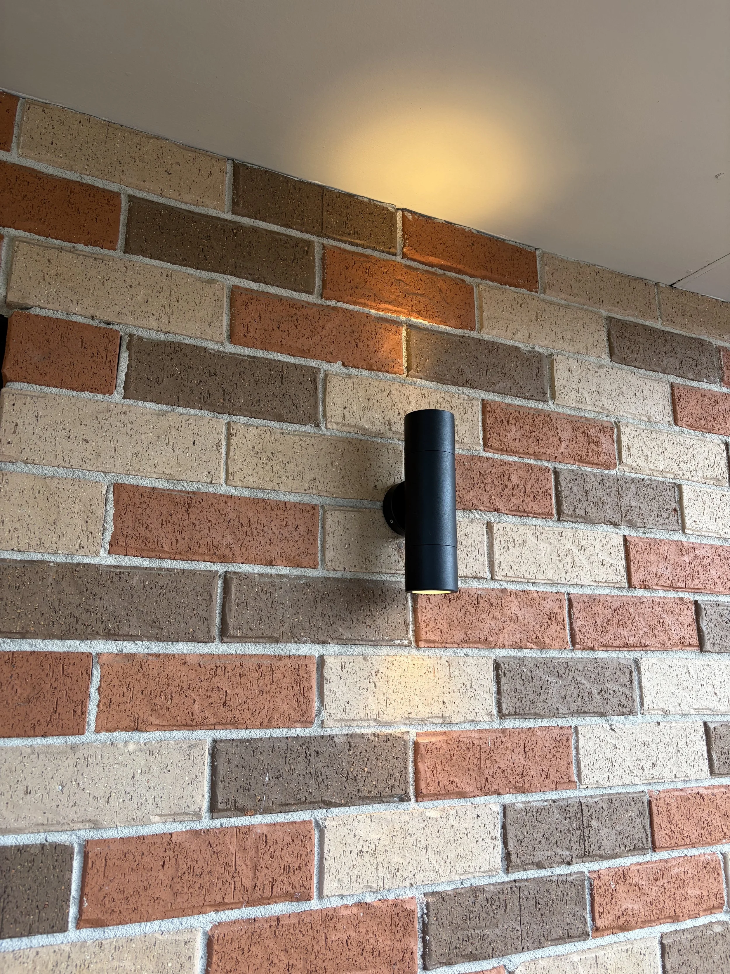 Black cylindrical outdoor wall light mounted on a brick wall, illuminating the ceiling above.