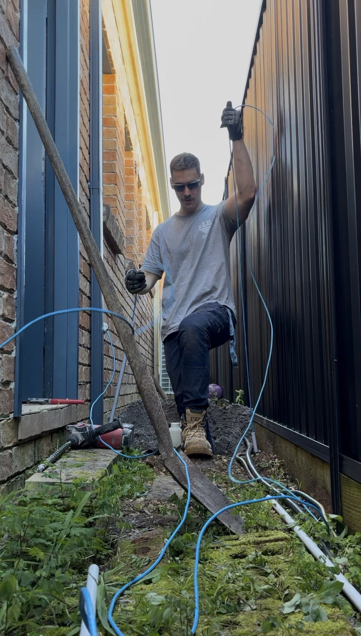 A man working on electrical wiring outside between a brick house and a black fence, wearing gloves, sunglasses, and work boots, surrounded by tools and cables.