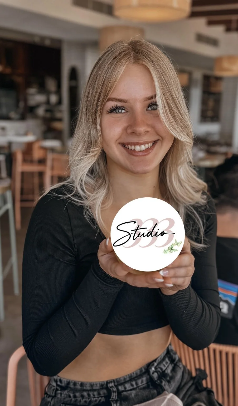 A smiling young woman with blonde hair holding a circular sign with the word "Studio333" and a small green vine illustration in a cozy, wooden interior space, possibly a cafe or studio.