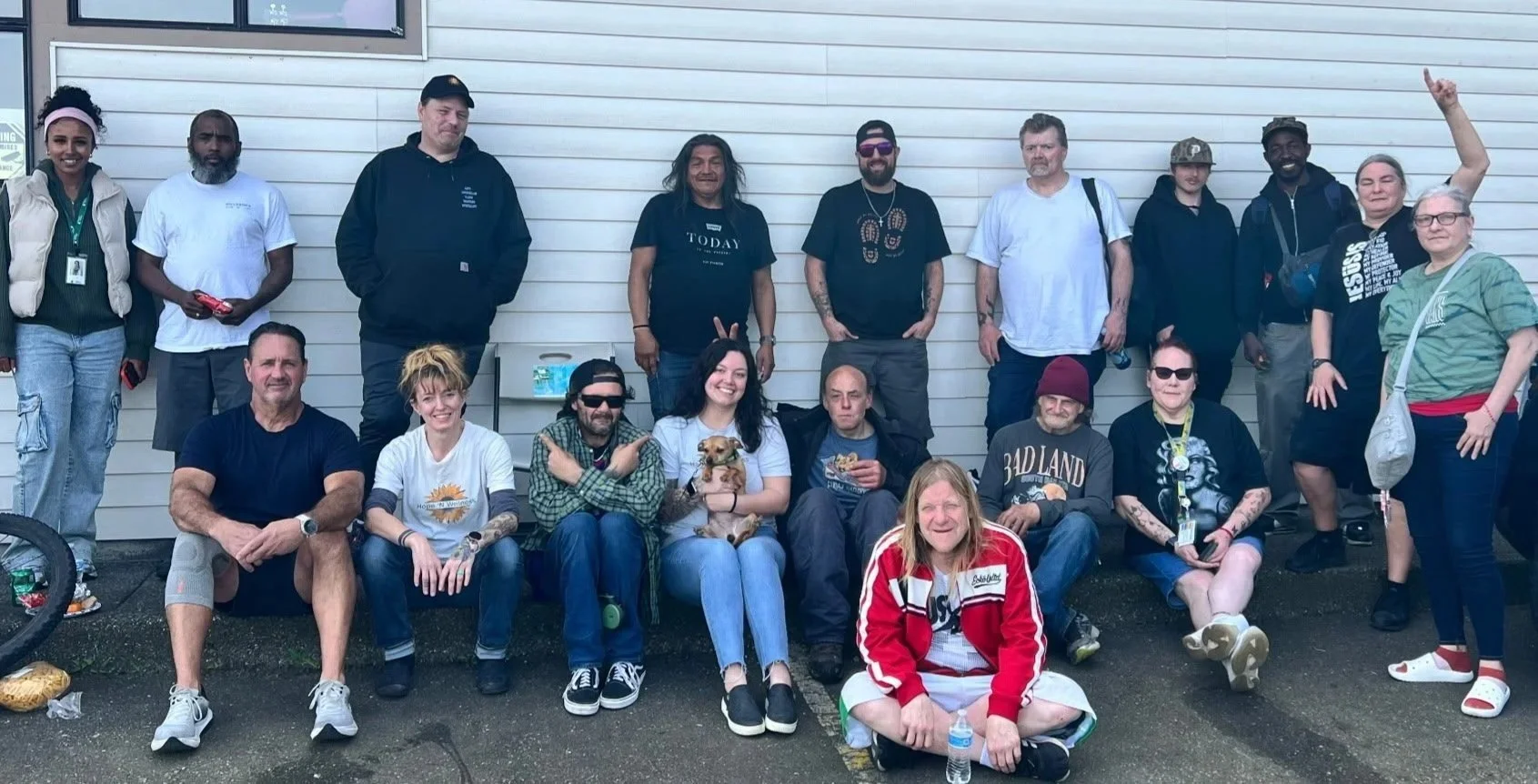 A group of 16 people and one dog pose outside against a beige building wall. Some people are sitting, some are standing, and one is reclining on the ground. The group appears cheerful, with some raising their hands or making peace signs.