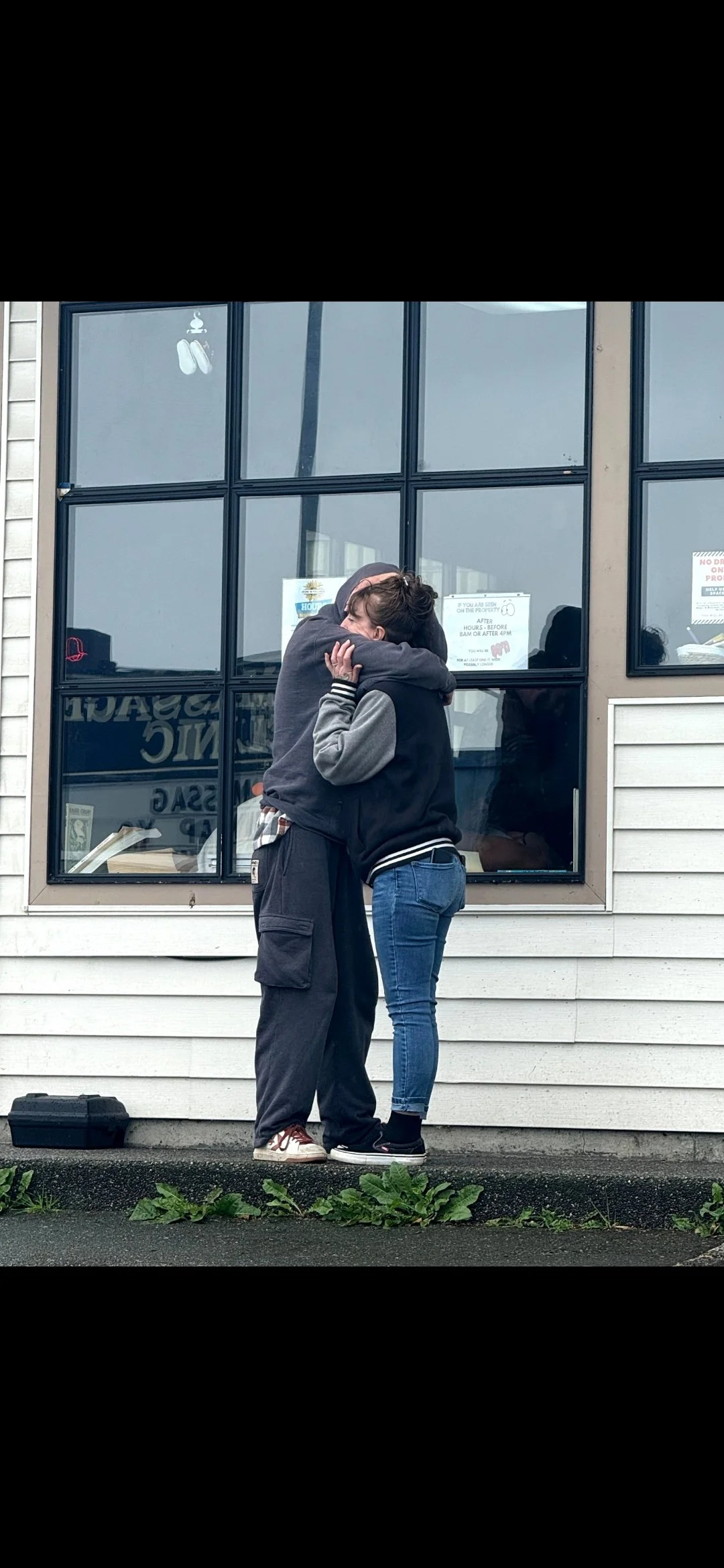 Two people hugging outside a building with large window panes, one wearing a gray and black hoodie, the other in a black jacket and blue jeans.