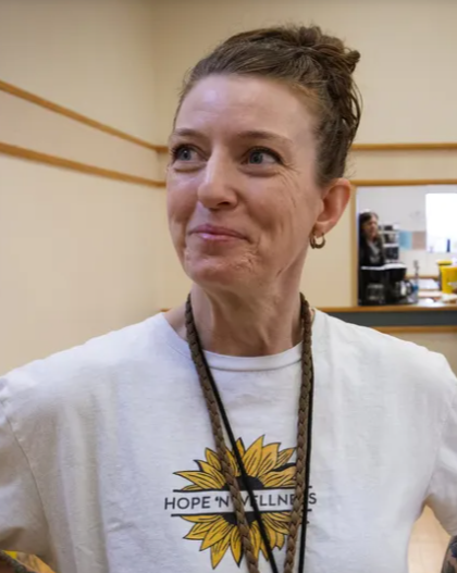 A woman smiling with her hair in a bun, wearing a white t-shirt with a yellow sunflower and the words "Hope & Wellness" printed on it, standing indoors near a counter.
