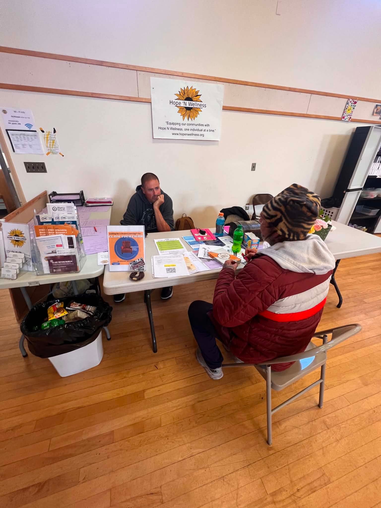 A man and a woman seated at a table in an indoor setting, possibly a community center or an office, with various documents, flyers, and beverages on the table. A sign on the wall behind them reads 'Hope N Wellness' and includes a sunflower logo.