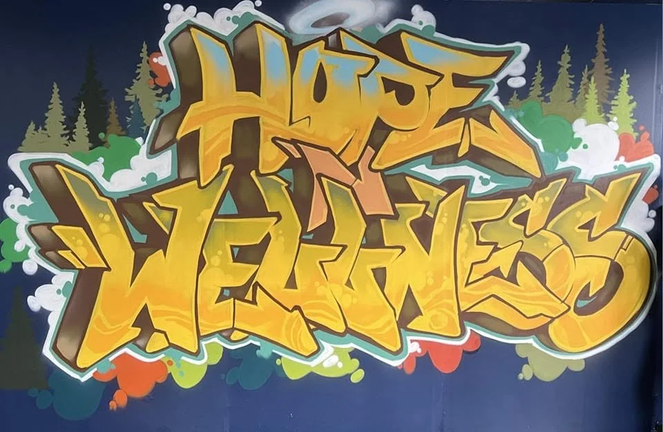 Colorful graffiti art with yellow, orange, and brown letters spelling "HOPE WARRIOR" against a dark background with green trees and clouds.