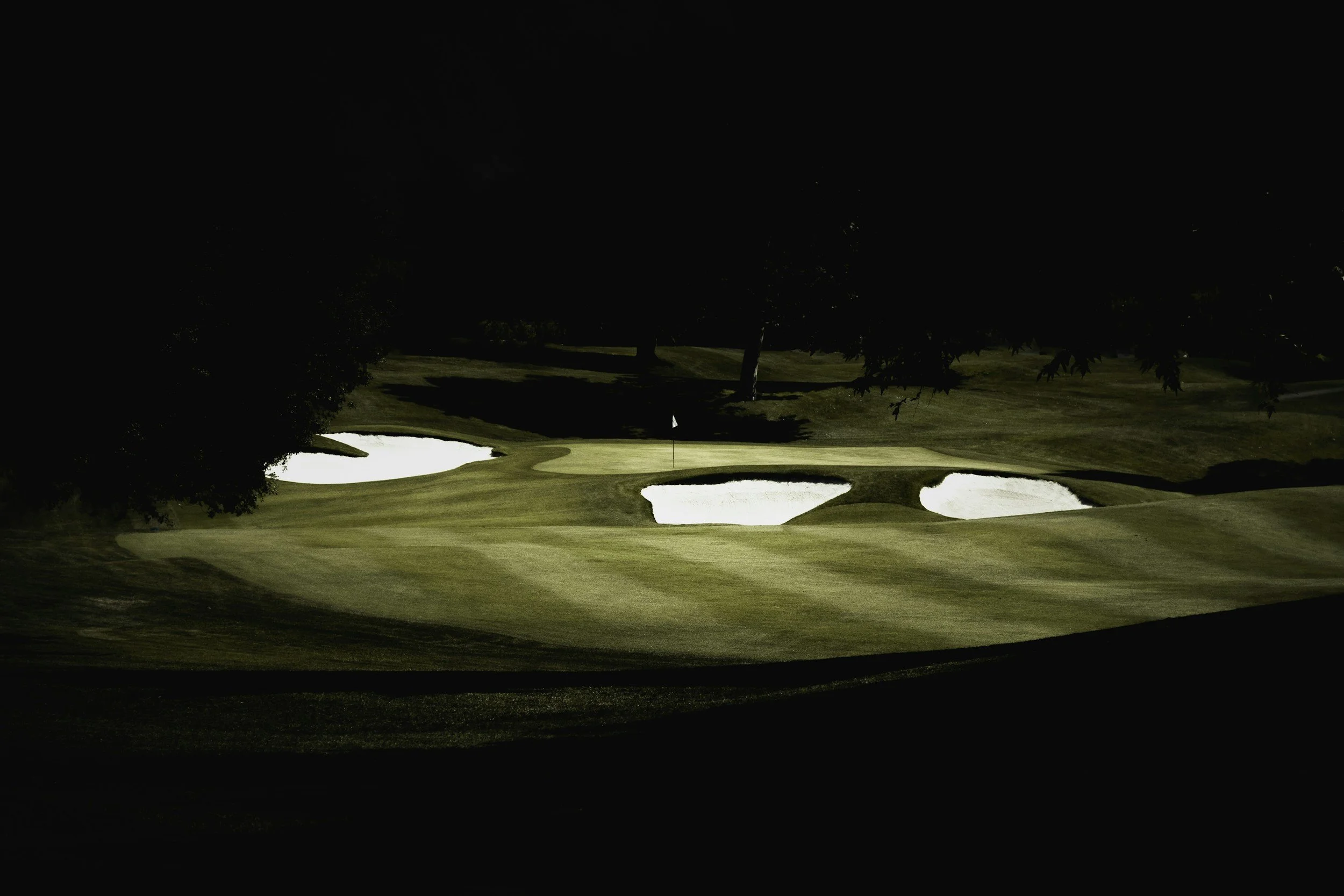 A golf course at night with a flag on the putting green and three sand bunkers surrounding it.