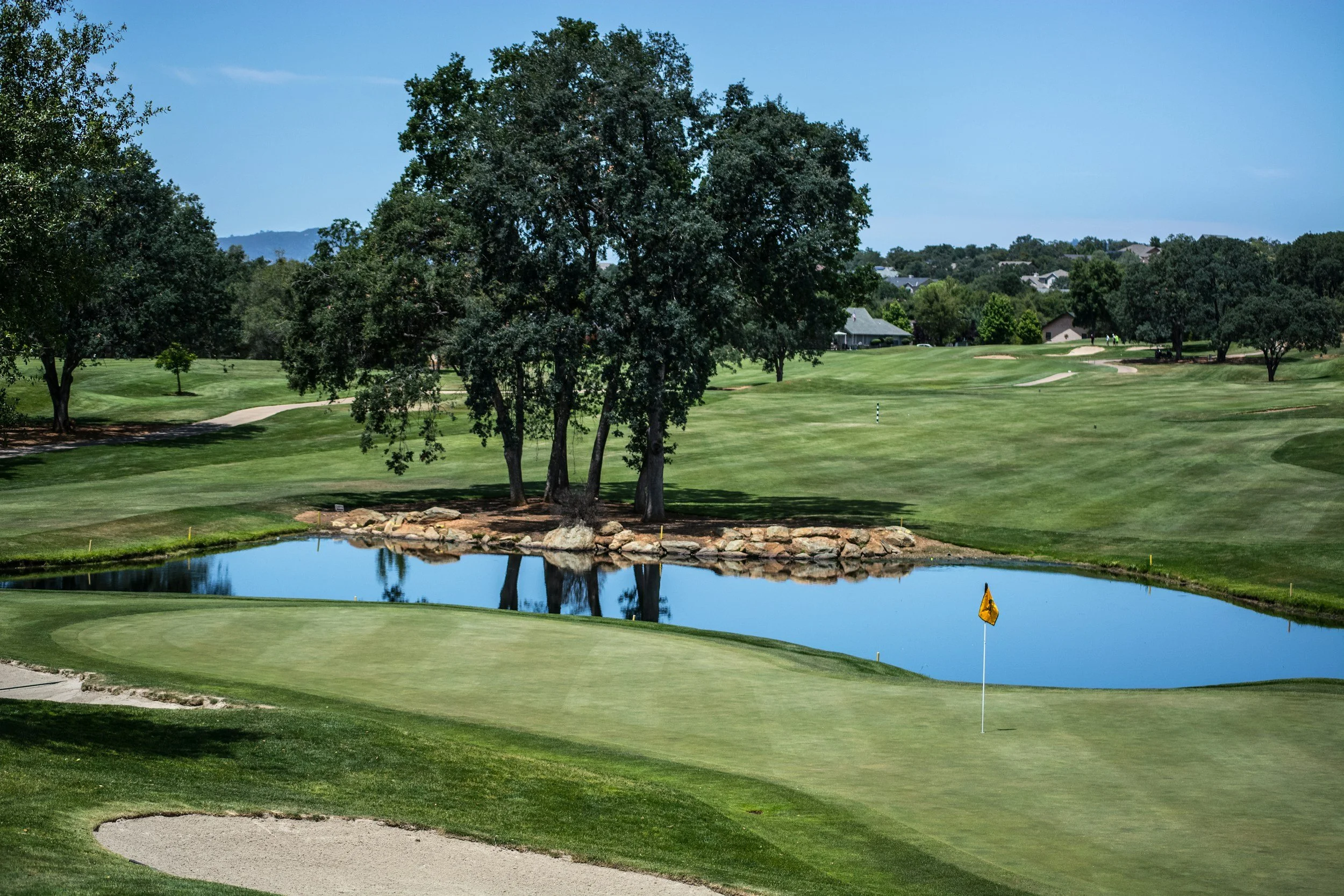 A golf course with a water hazard, a yellow flag on the green, and trees in the background under a clear blue sky.