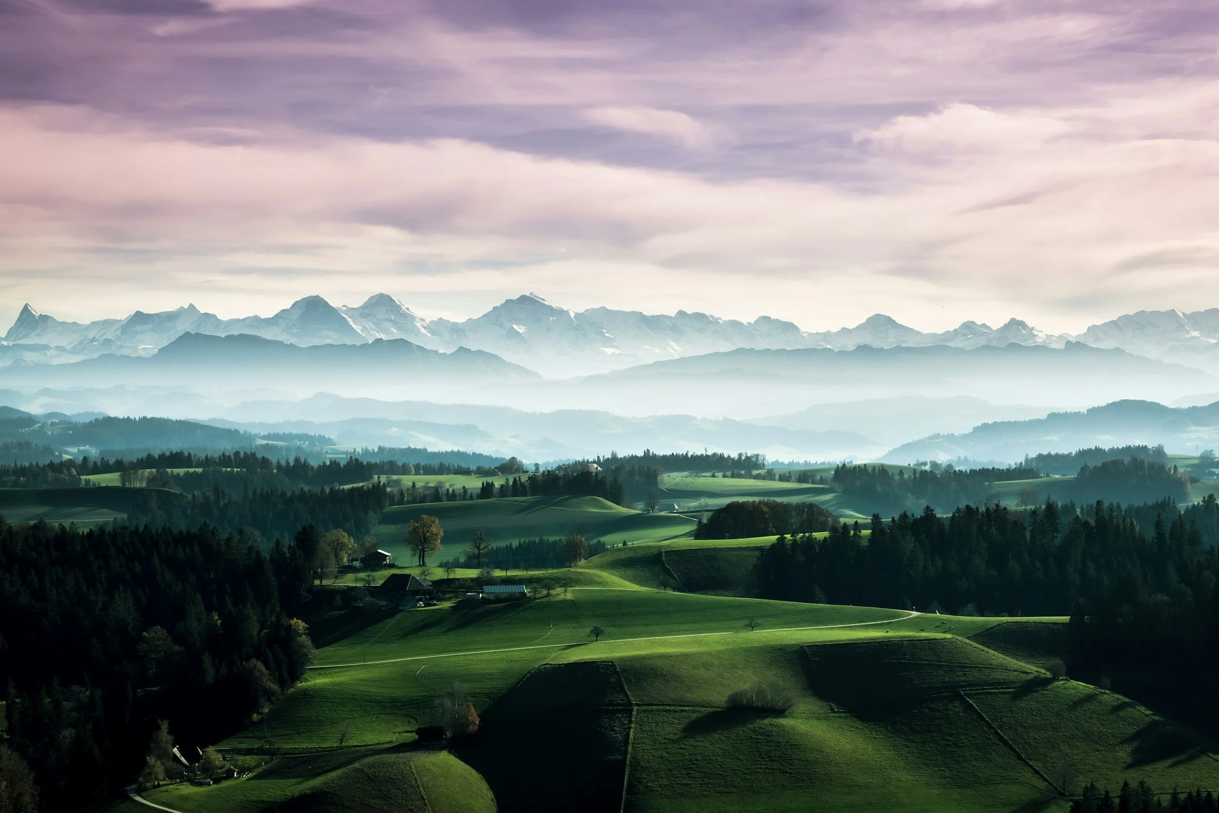 Scenic landscape of rolling green hills and farmland in the foreground, with a misty mountain range in the background under a cloudy sky.