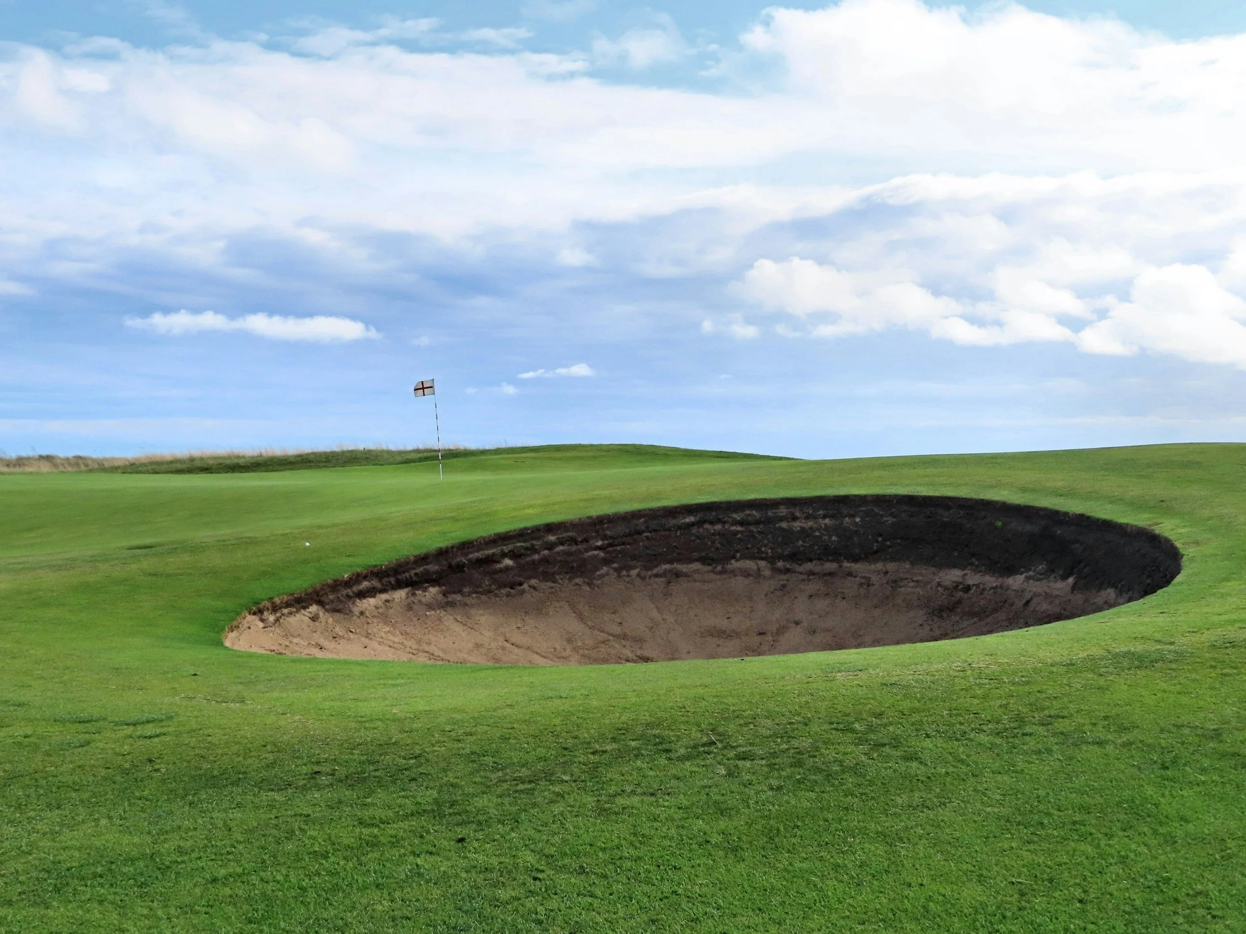 A golf course green with a sand bunker in the foreground and a flagstick with a red cross on a white flag on the green in the background under a partly cloudy sky.
