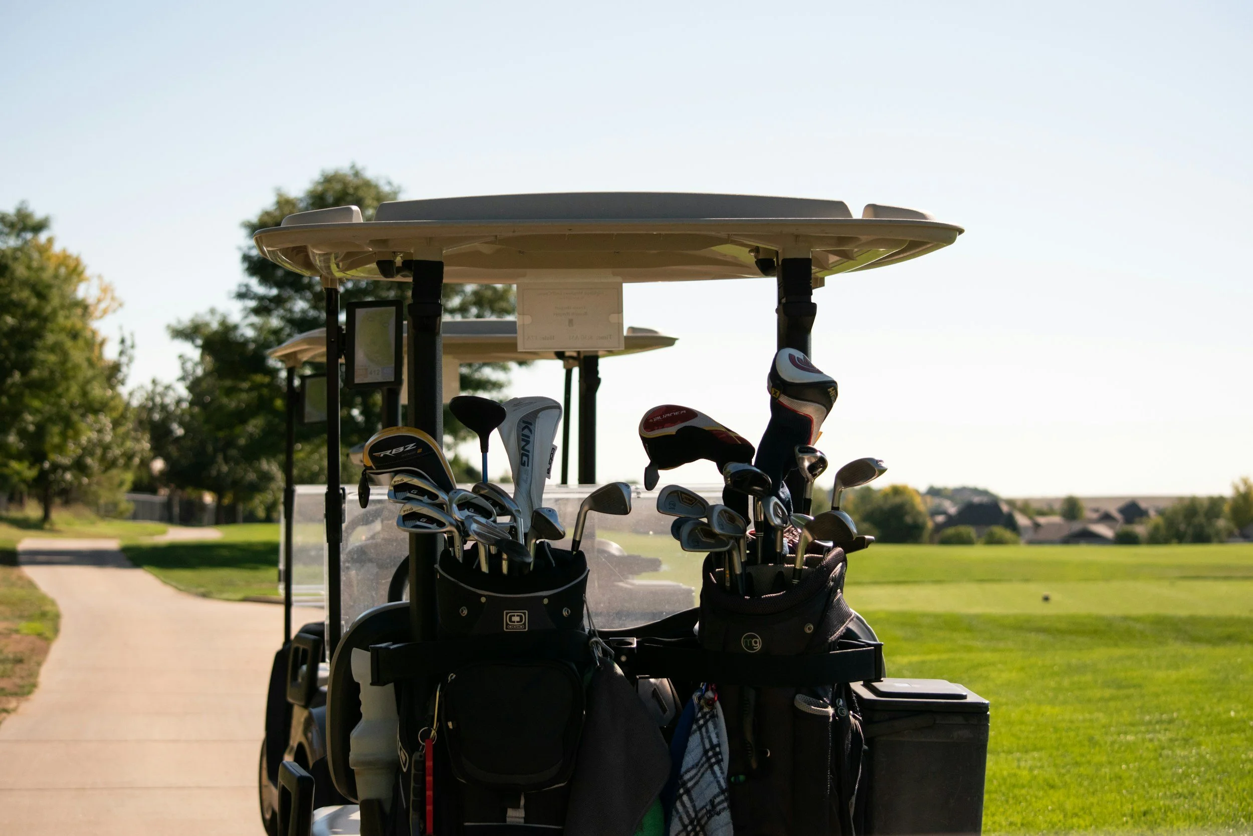 Golf cart with golf bags and clubs parked on a golf course trail in sunlight, with trees and houses in the background.