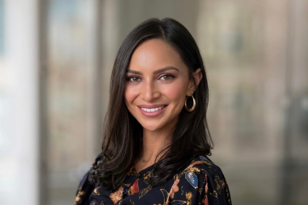 A woman with long, dark hair smiling, wearing a floral-patterned top and gold hoop earrings, with a blurred indoor background.