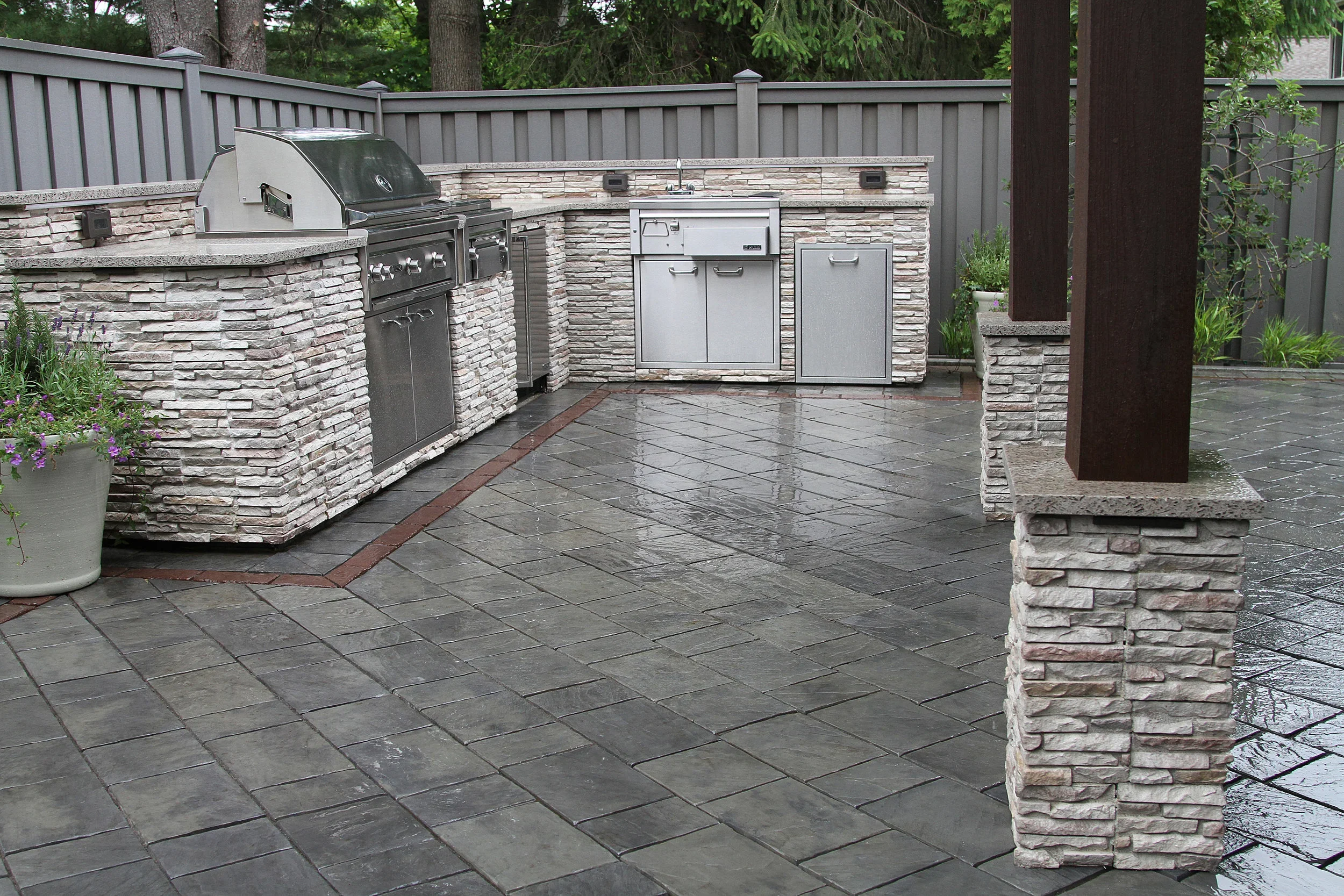 Outdoor kitchen with built-in grill, sink, and storage, surrounded by a stone counter and a gray privacy fence, with wet tiled flooring and a potted plant with purple flowers.