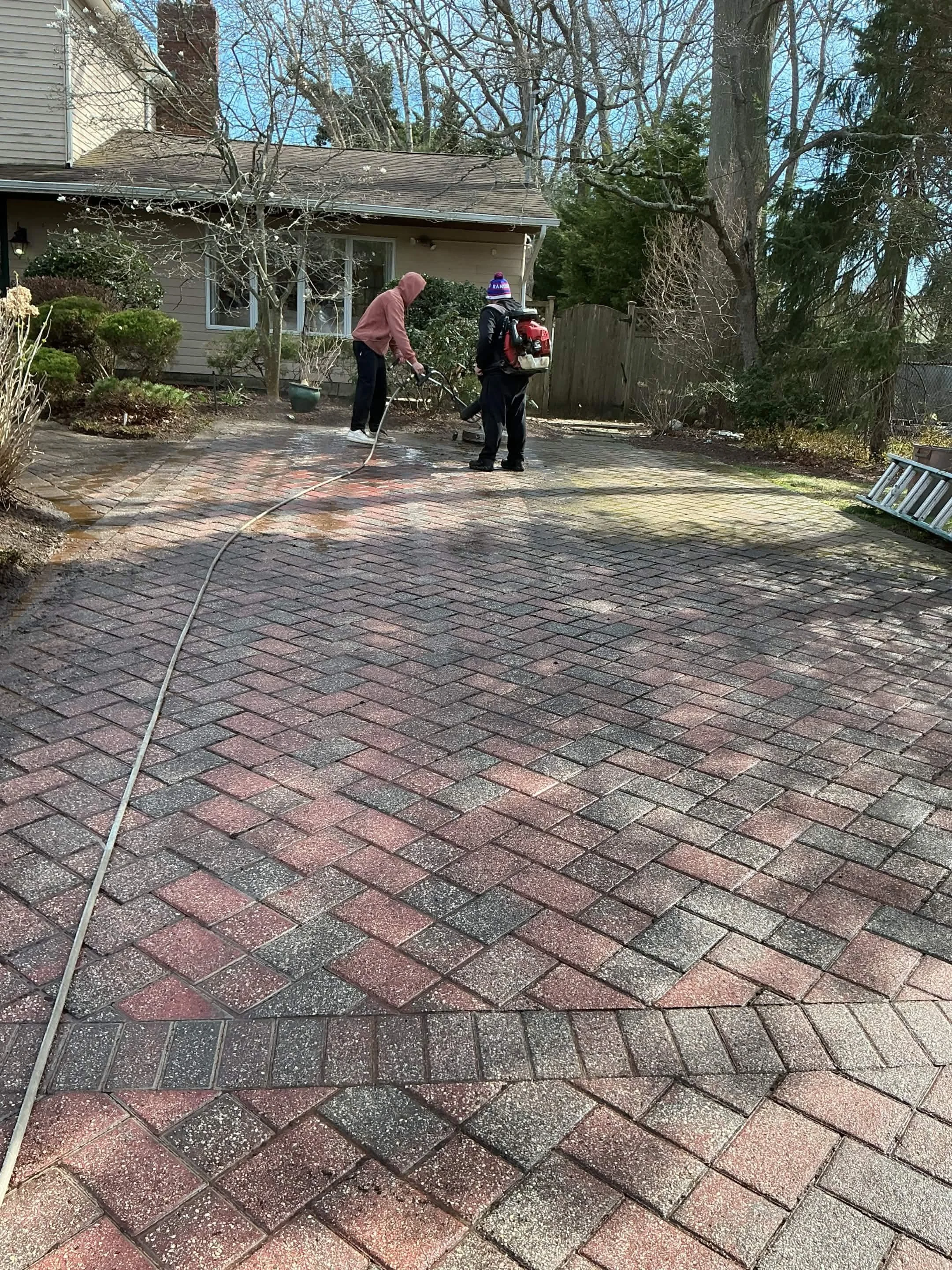 Two people power-washing a brick patio in a backyard with trees and shrubs.