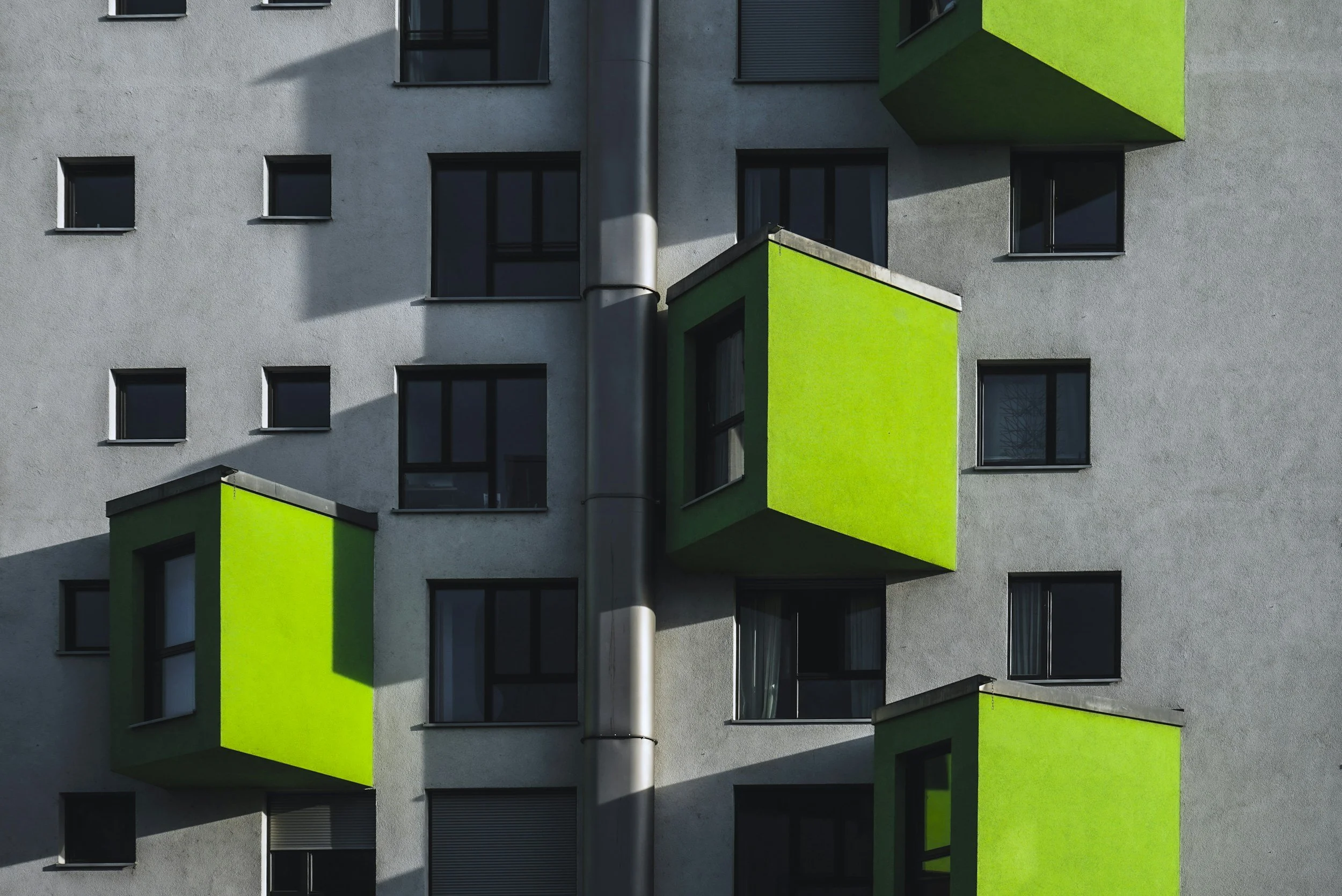 Modern apartment building with gray facade and green cubic balconies, some windows with black frames, and a vertical metal pipe in the center. Shadows cast across the building.