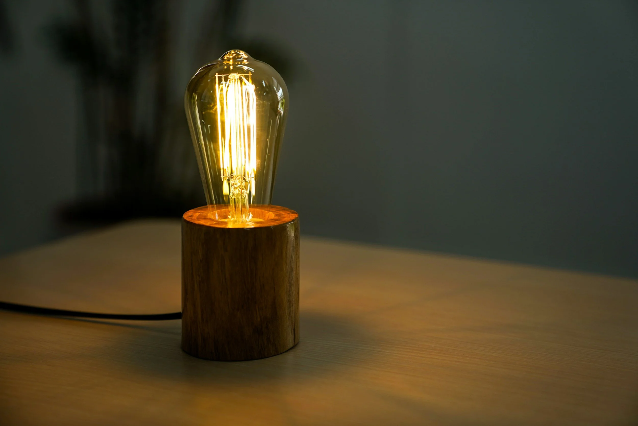 A table lamp with a vintage-style filament bulb on a wooden base, casting a warm glow in a dim room.