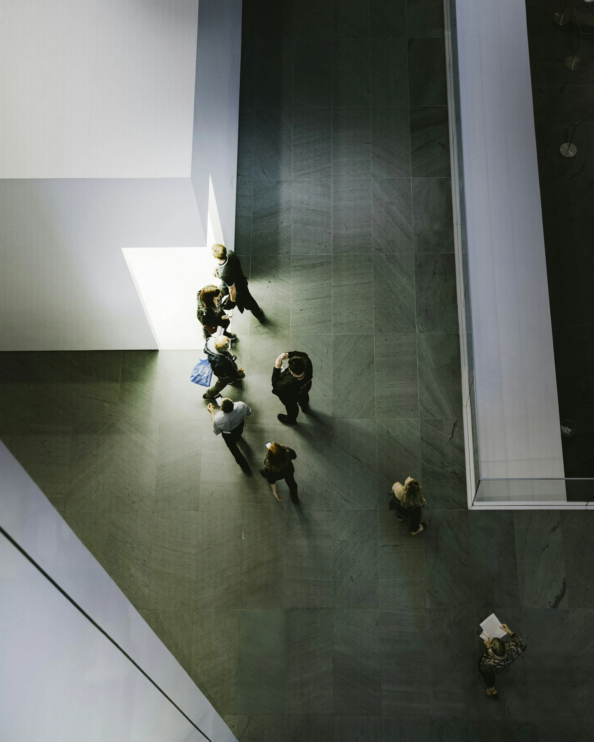 Top-down view of people standing and walking in a modern indoor space with gray tiled flooring and white walls, with sunlight casting shadows.