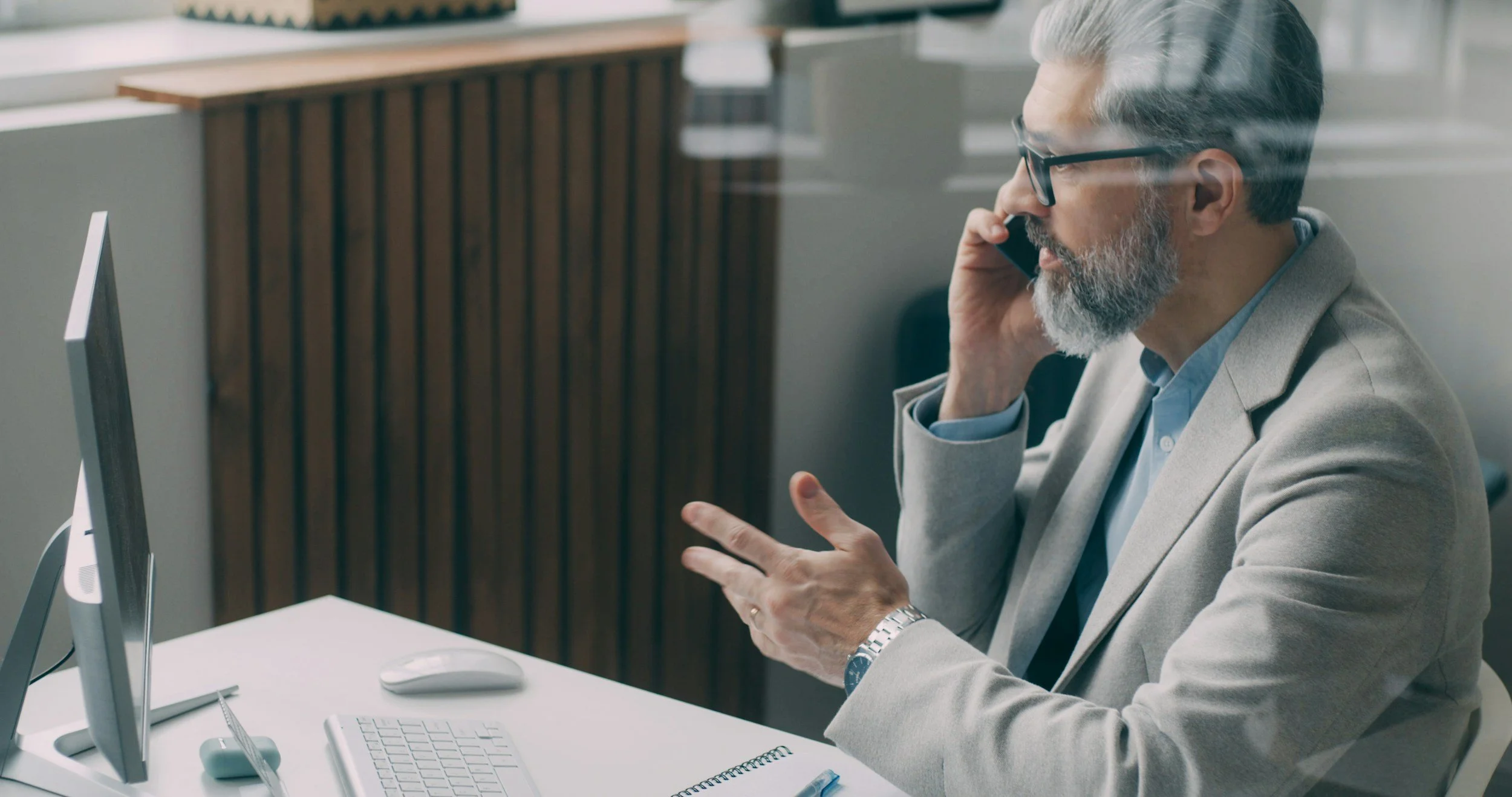 An older man with gray hair and beard talking on his smartphone in an office, sitting at a desk with a computer, keyboard, mouse, and notepad.
