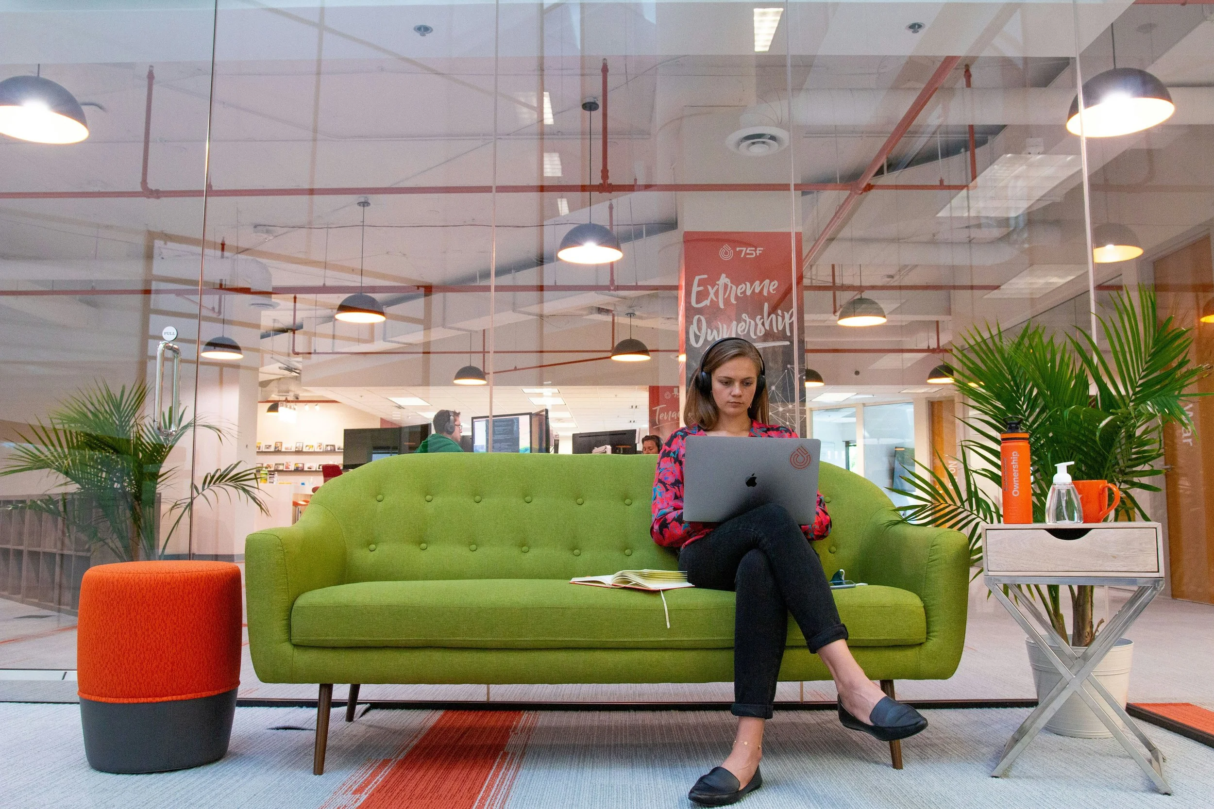 A woman sitting on a green sofa in an office lobby, working on her laptop with headphones on, with a book open on her lap, surrounded by plants and a small table with drink bottles and a hand sanitizer.