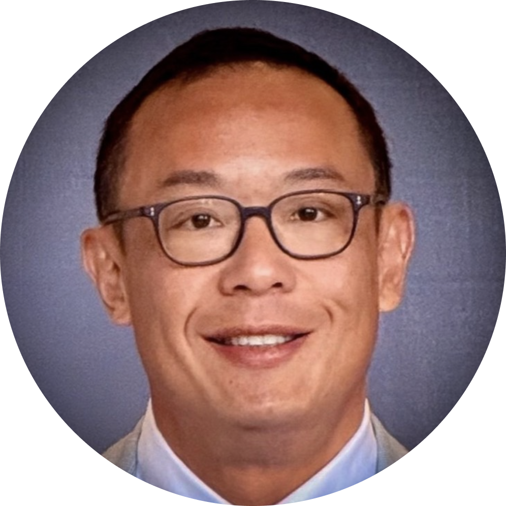 A man with glasses and short dark hair smiling, wearing a light-colored suit jacket and a collared shirt, against a blue background.