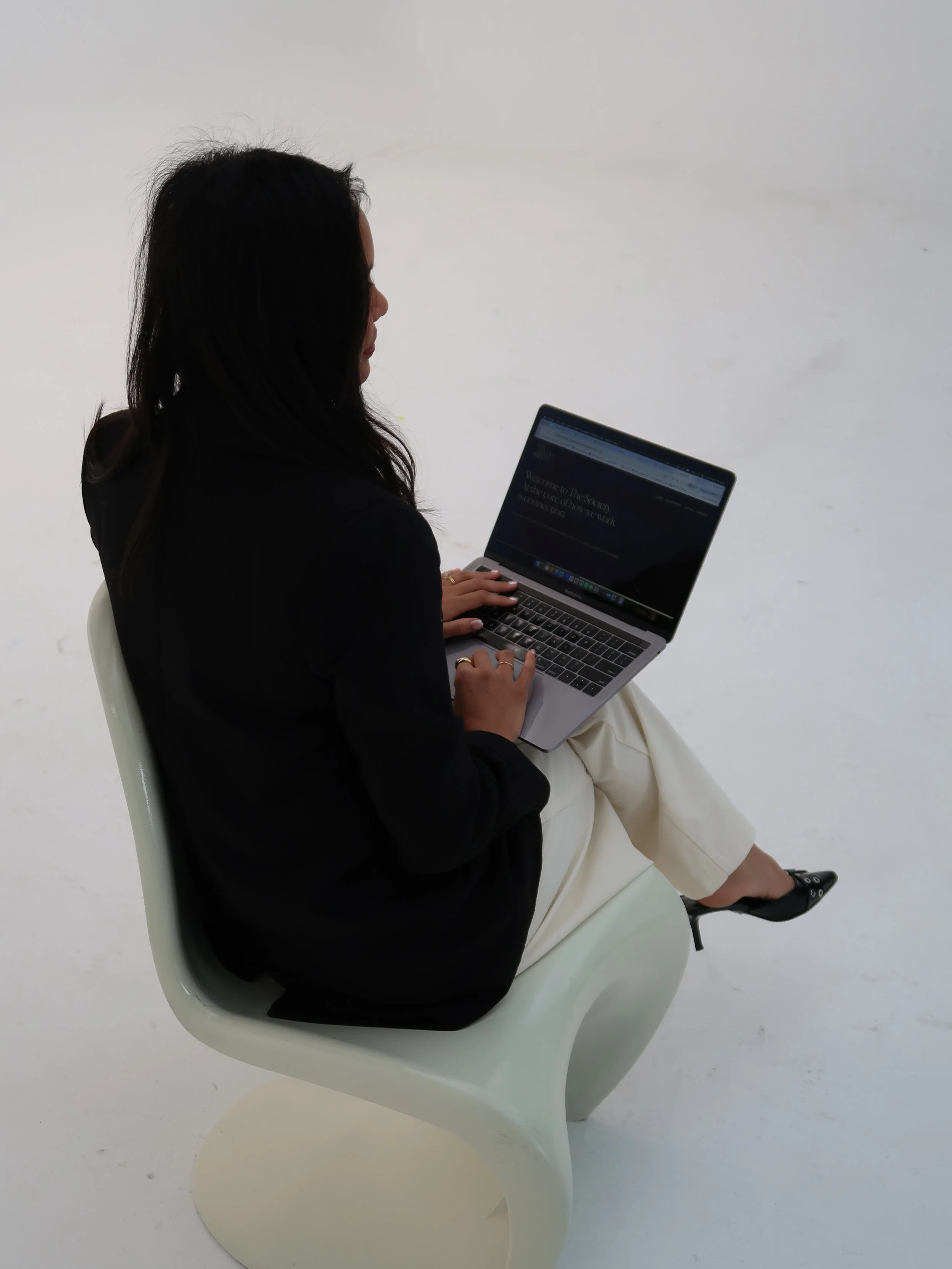 A woman with dark hair sitting on a white chair, working on a laptop, in a minimalist setting with a plain white background.