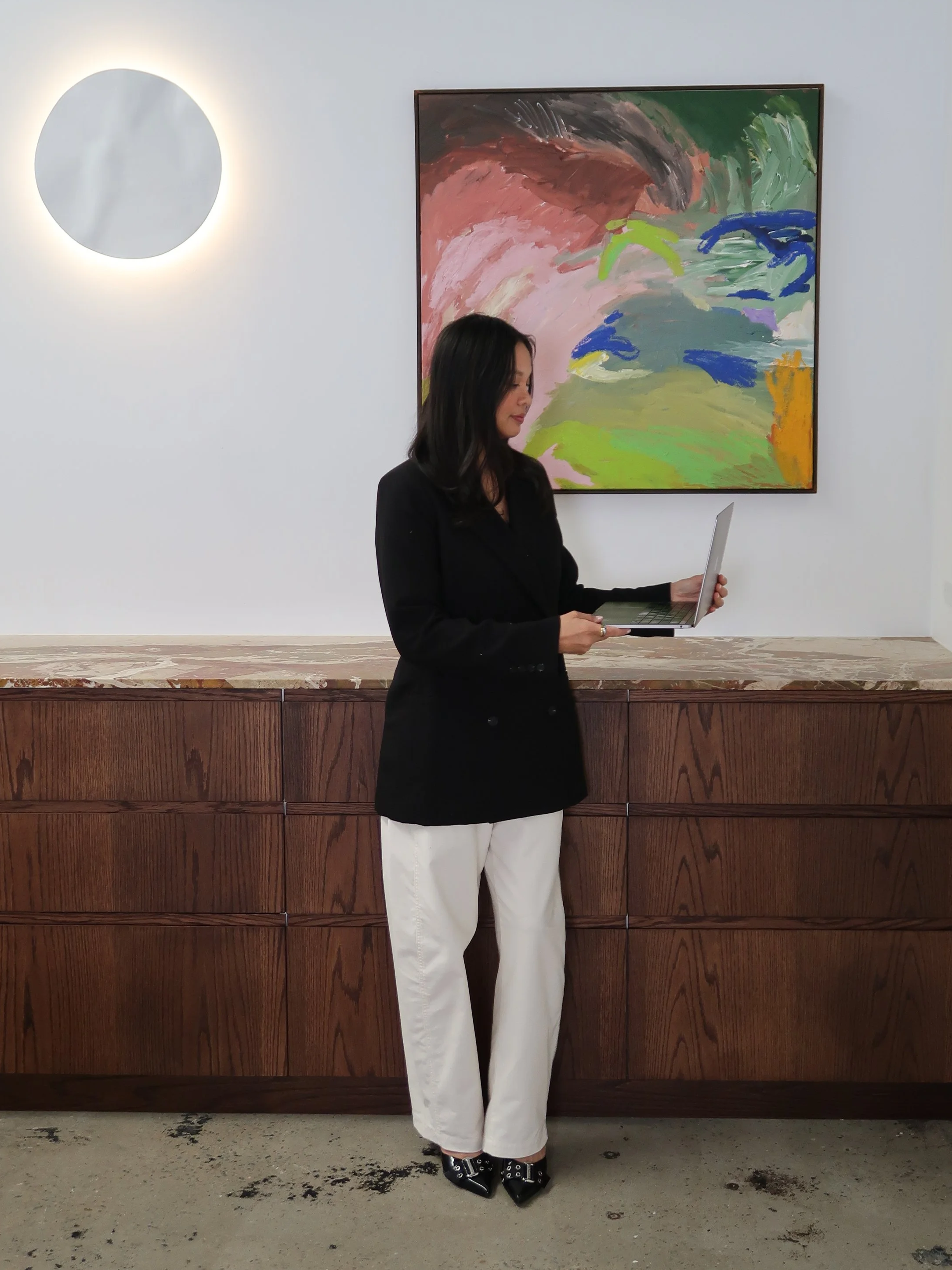 Woman standing in front of a white wall with colorful abstract painting, holding a laptop, wearing a black blazer, white wide-leg pants, and black pointed shoes with buckles.