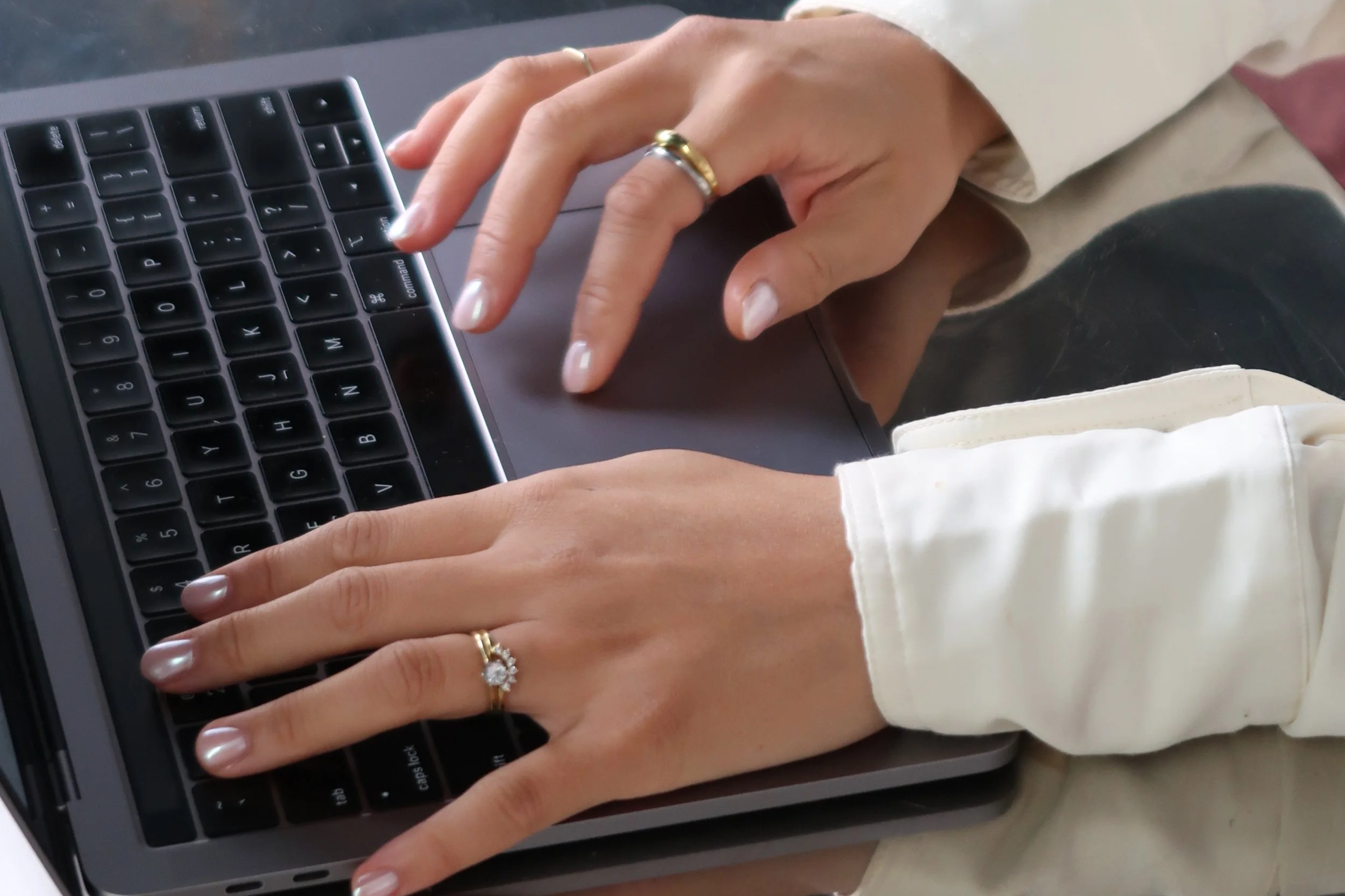 Close-up of a person's hands typing on a laptop keyboard. The person is wearing a gold ring with a floral design on their ring finger and another gold ring on their middle finger. They are dressed in a light-colored shirt or blouse with a long sleeve visible.