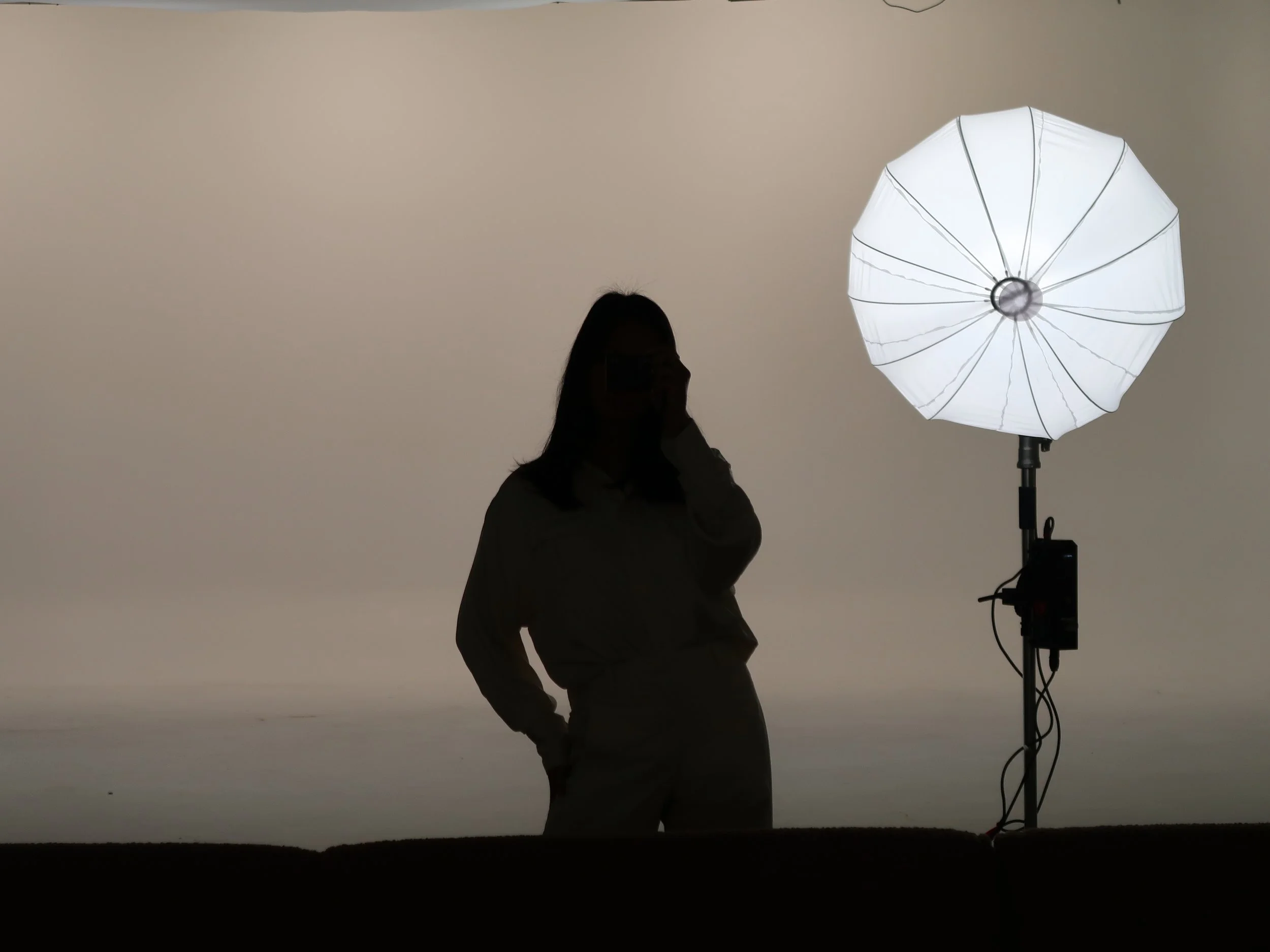 A person with long hair, wearing glasses and a light-colored shirt, stands next to a large, white photography umbrella light on a stand in a photo studio with a plain background.