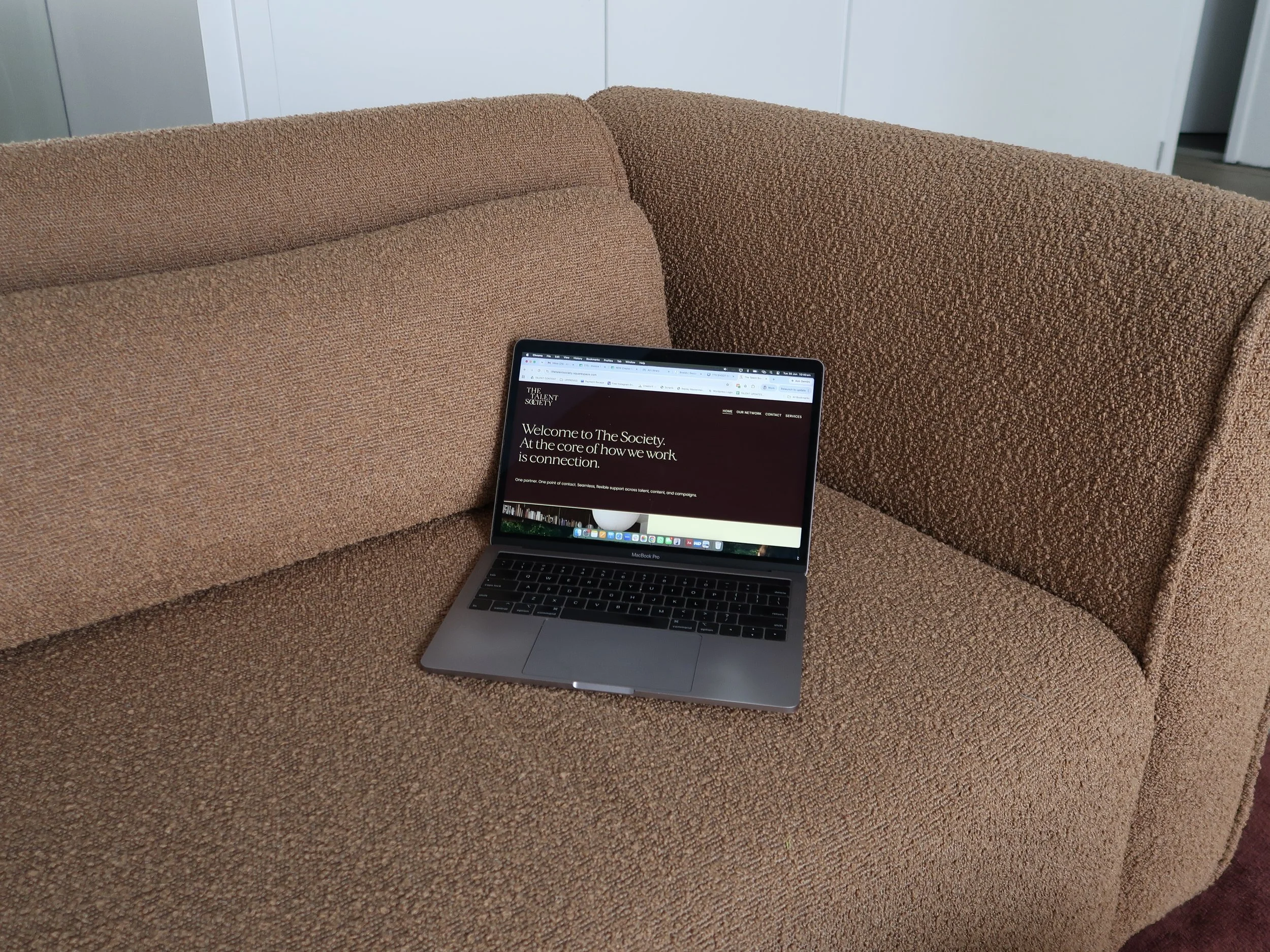 A MacBook Pro laptop on a beige textured sofa with a web page displaying 'Welcome to The Society' open on the screen. The sofa has a brown fabric and is positioned in a modern office or lounge area with white walls and cabinets.