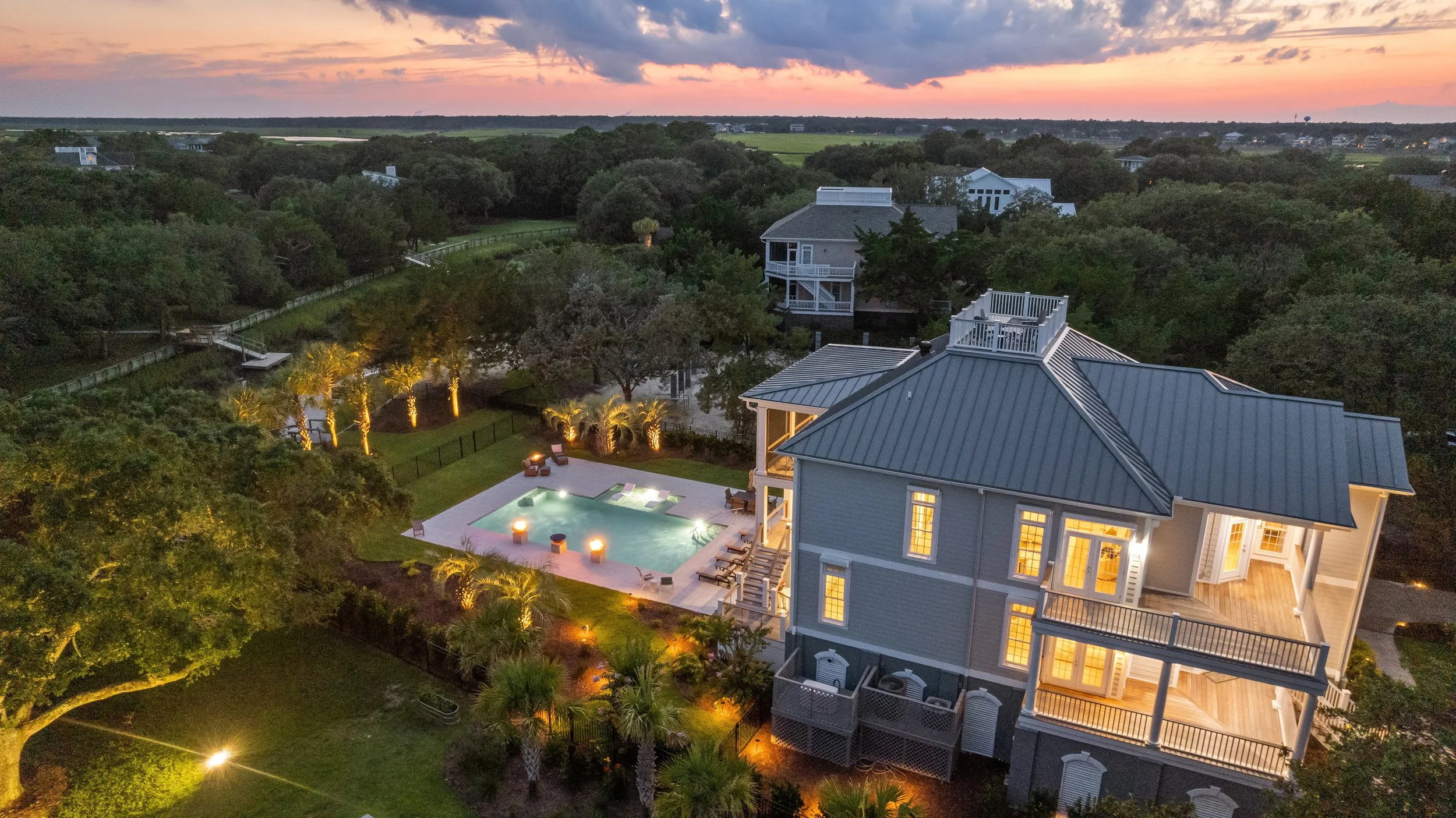 Aerial view of a large house with a metal roof, balcony, and outdoor lighting at dusk, surrounded by lush trees and a backyard swimming pool with lounge chairs. In the background, there are more houses, open fields, and a colorful sunset sky.