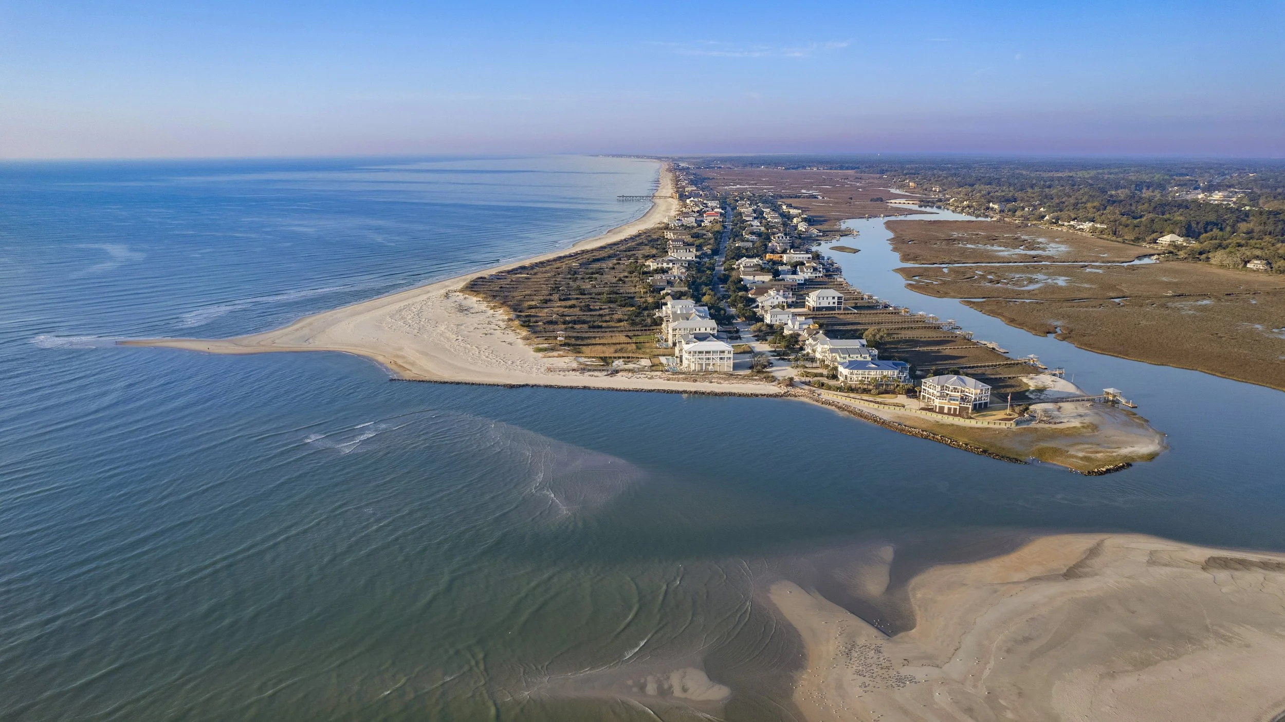 Aerial view of a coastal residential area with houses and buildings along a long, narrow stretch of beach and a river or creek running parallel to the shoreline.