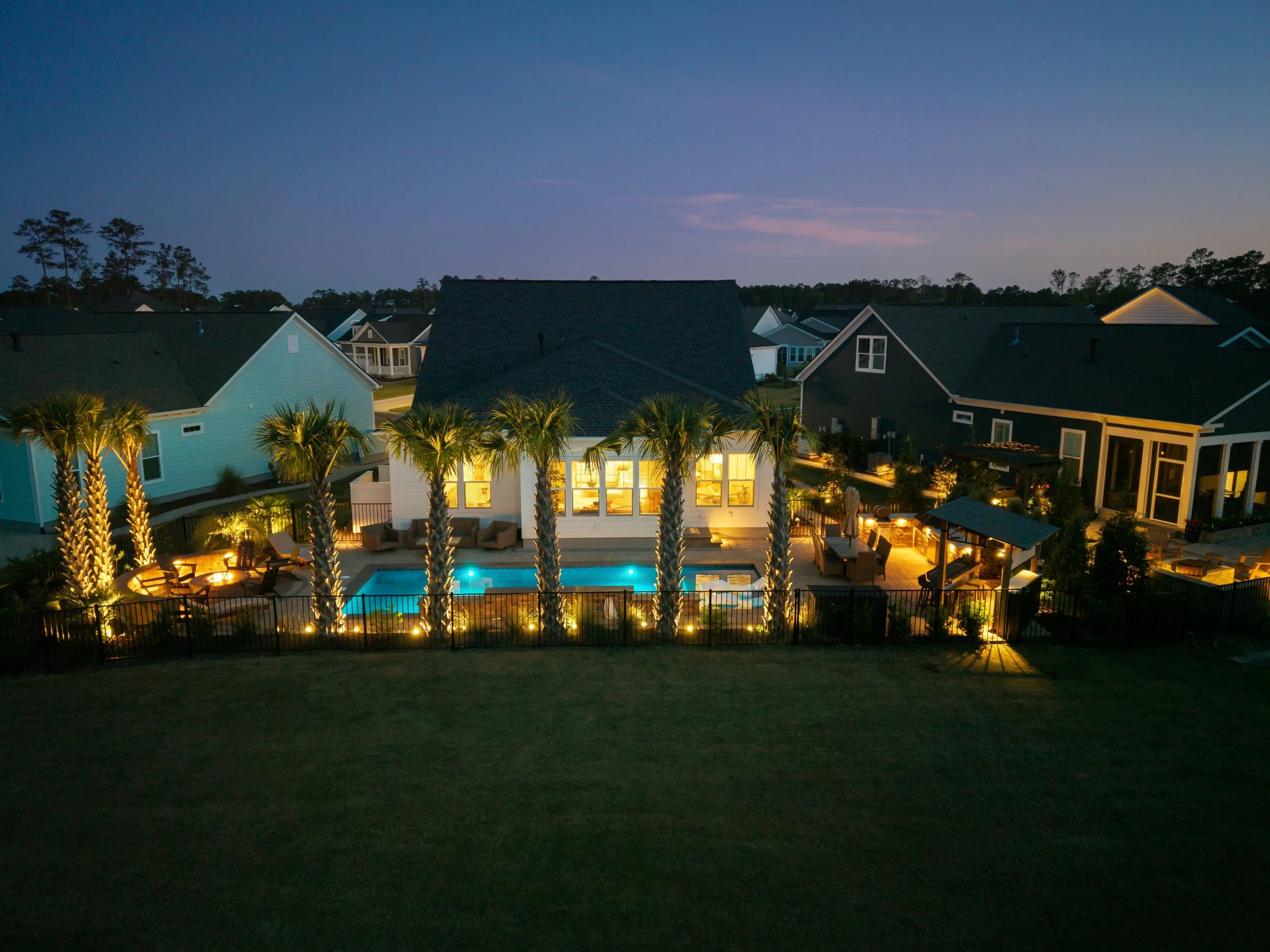 A backyard at dusk with a lit swimming pool, patio furniture, palm trees, and houses with illuminated windows.