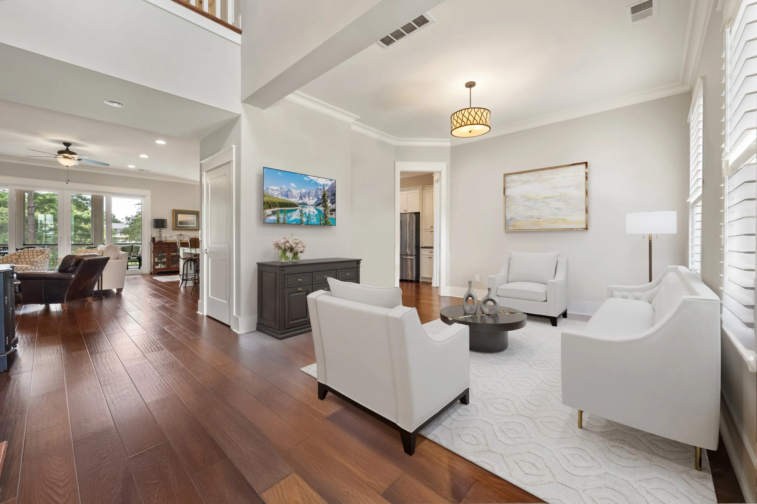 Living room with white walls, wooden floors, white and neutral-colored furniture, and artwork on the walls. Television on the wall, ceiling fans, large windows with shutters, and decorative vases on a black side table.