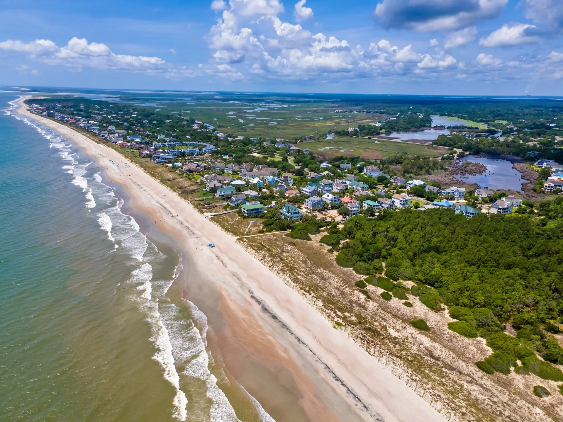Aerial view of a coastal area with a sandy beach, waves, residential houses, and green vegetation under a partly cloudy sky.