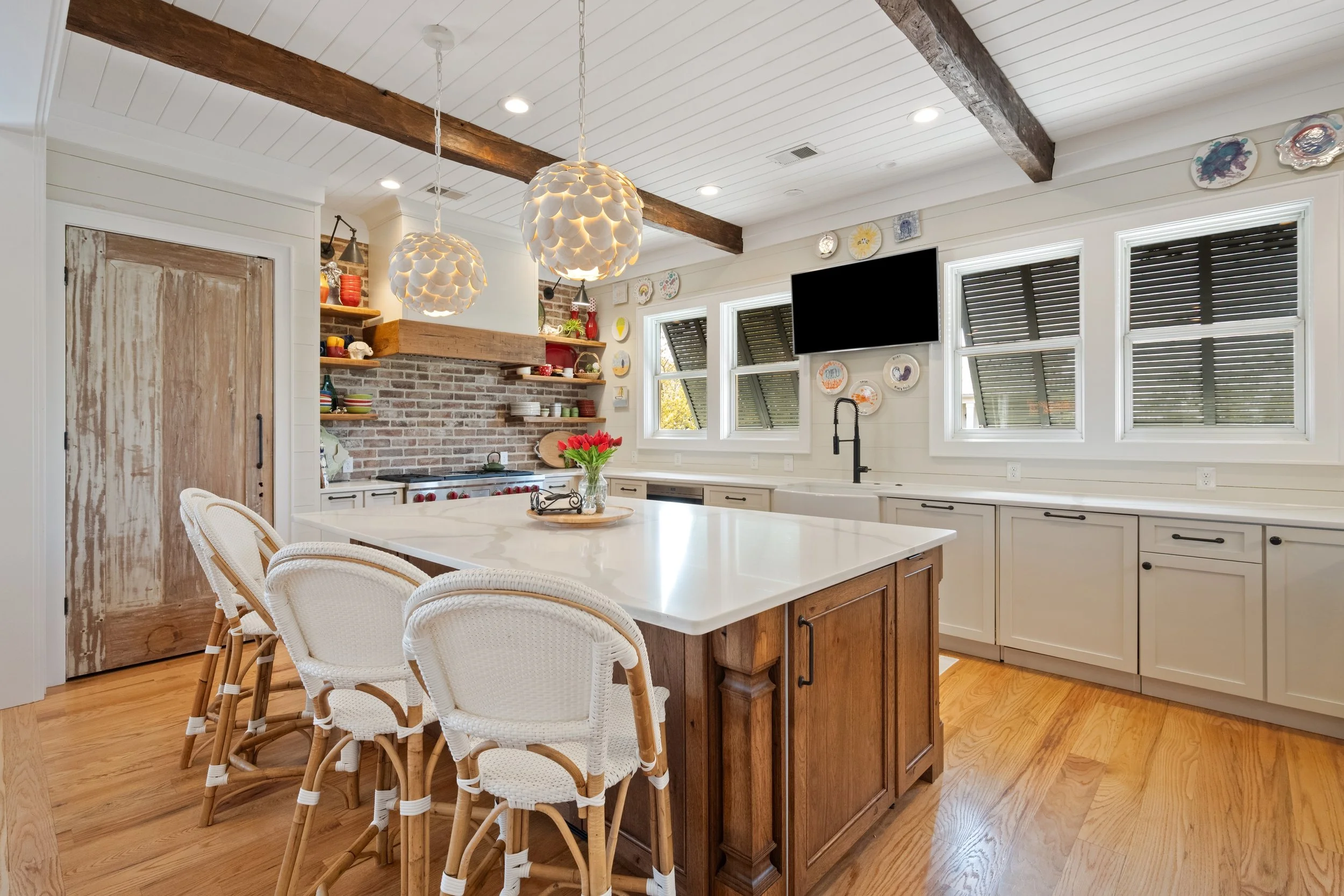 A modern kitchen with a large white island, four white wicker chairs with wooden legs, wood flooring, a brick backsplash, open wooden shelves with dishes and decor, a large wooden beam on the ceiling, and four windows with louvered blinds.