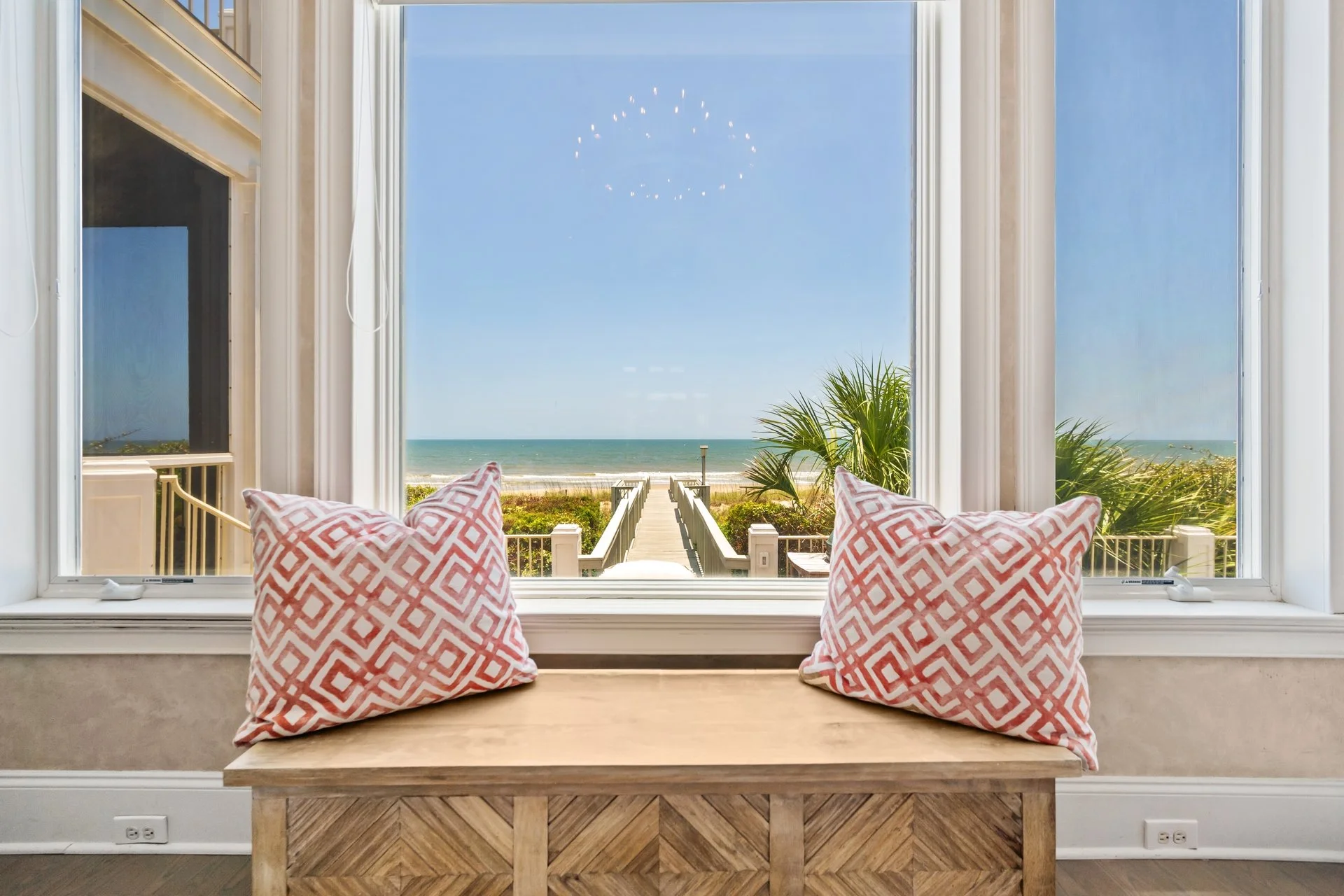 Beach view with a sandy pathway leading to the ocean, seen through a large window with two pink and white patterned pillows on a wooden bench inside.