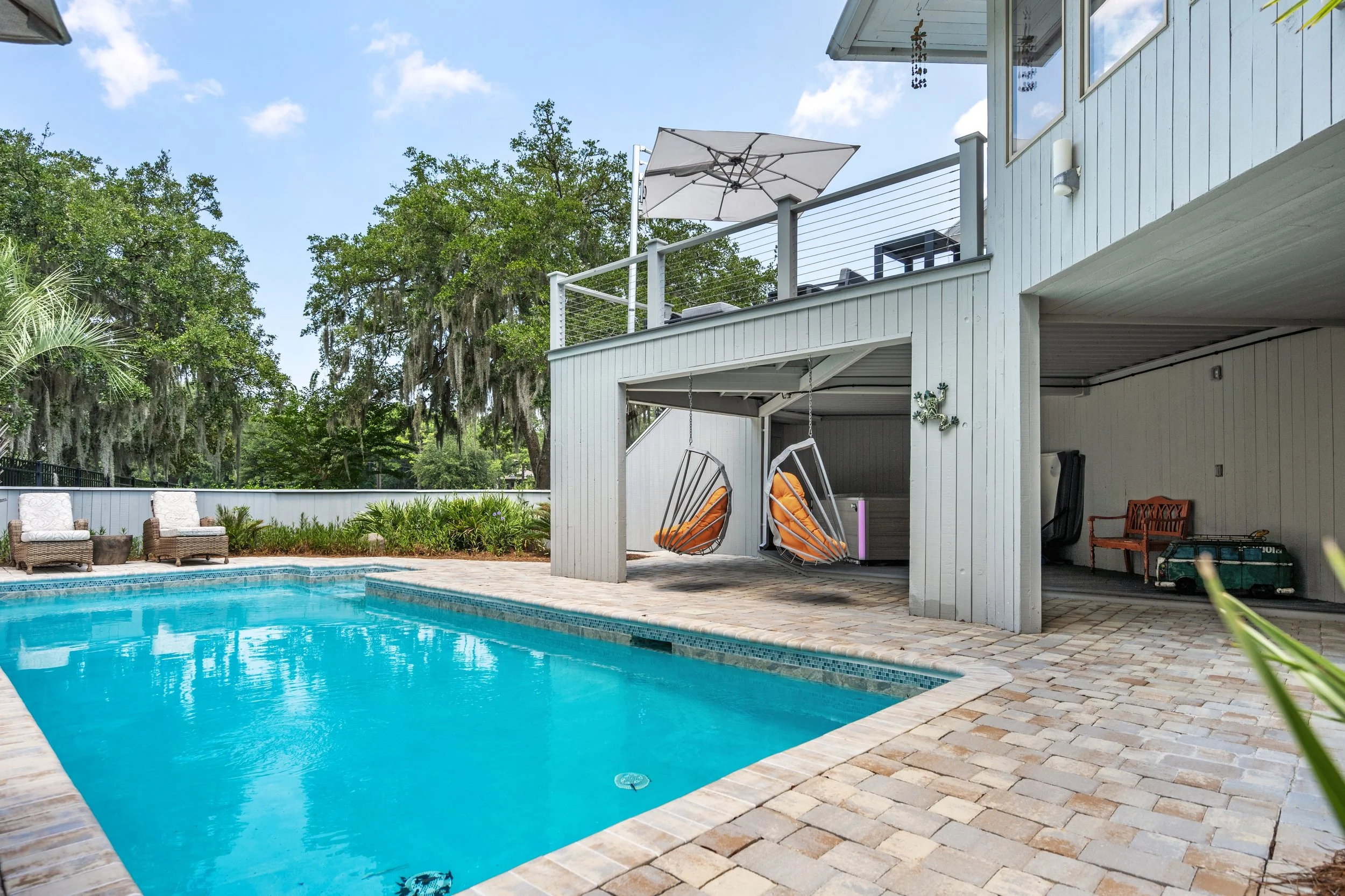A backyard pool area with lounge chairs on a brick deck, a white house with a covered patio, hanging swing chairs, and lush green trees in the background.