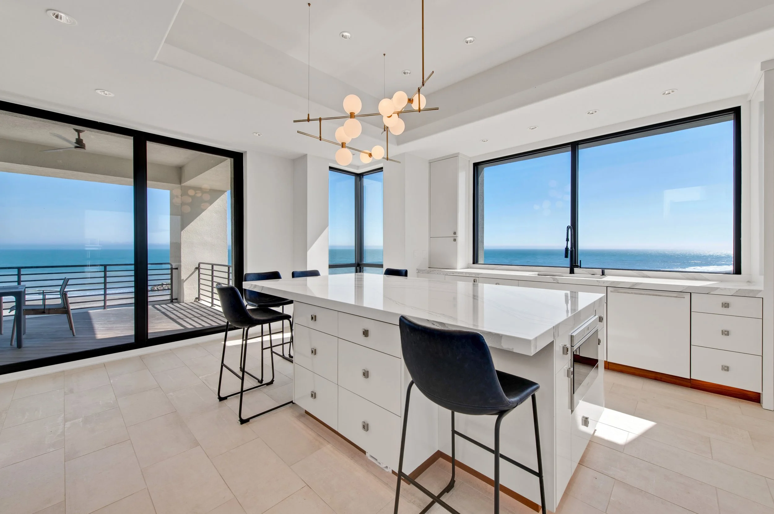 Modern kitchen with large windows overlooking the ocean, white cabinets, marble island, black chairs, and a pendant light fixture.