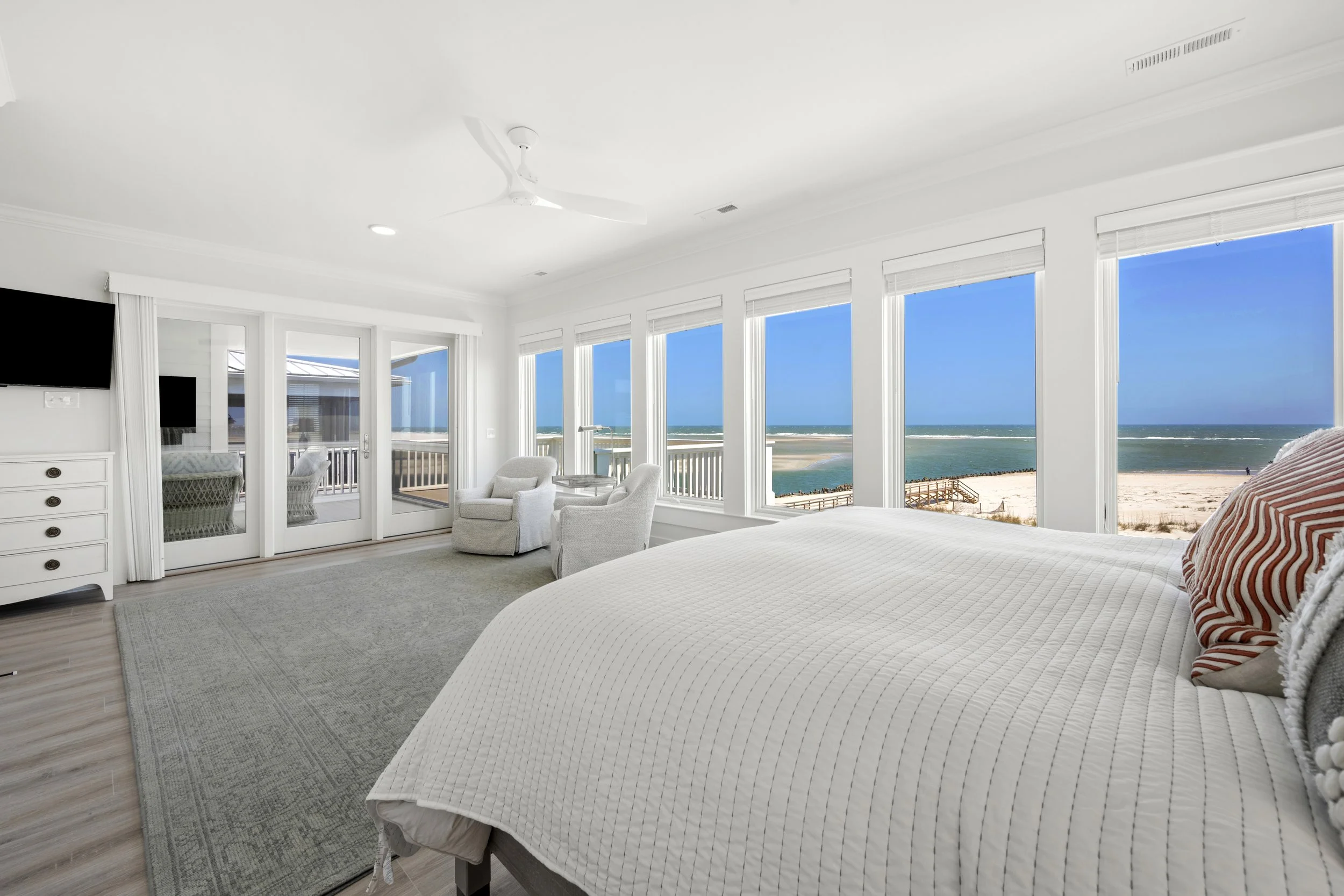 A bright, airy bedroom with large windows overlooking a beach and ocean, furnished with a white bed, white armchairs, and a white dresser.
