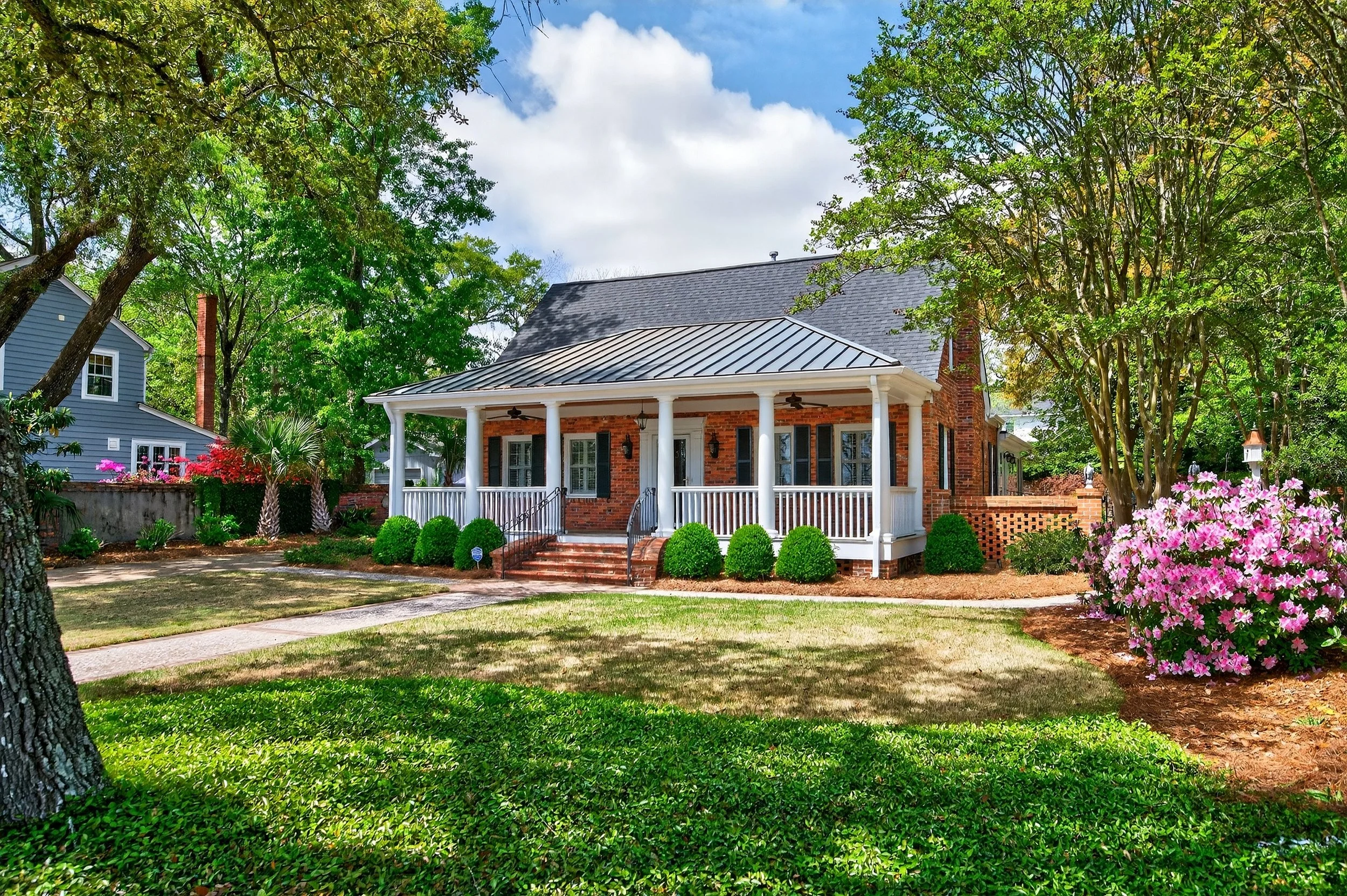 Front view of a brick house with white porch columns, surrounded by green trees and pink flowering bushes, under a partly cloudy sky.