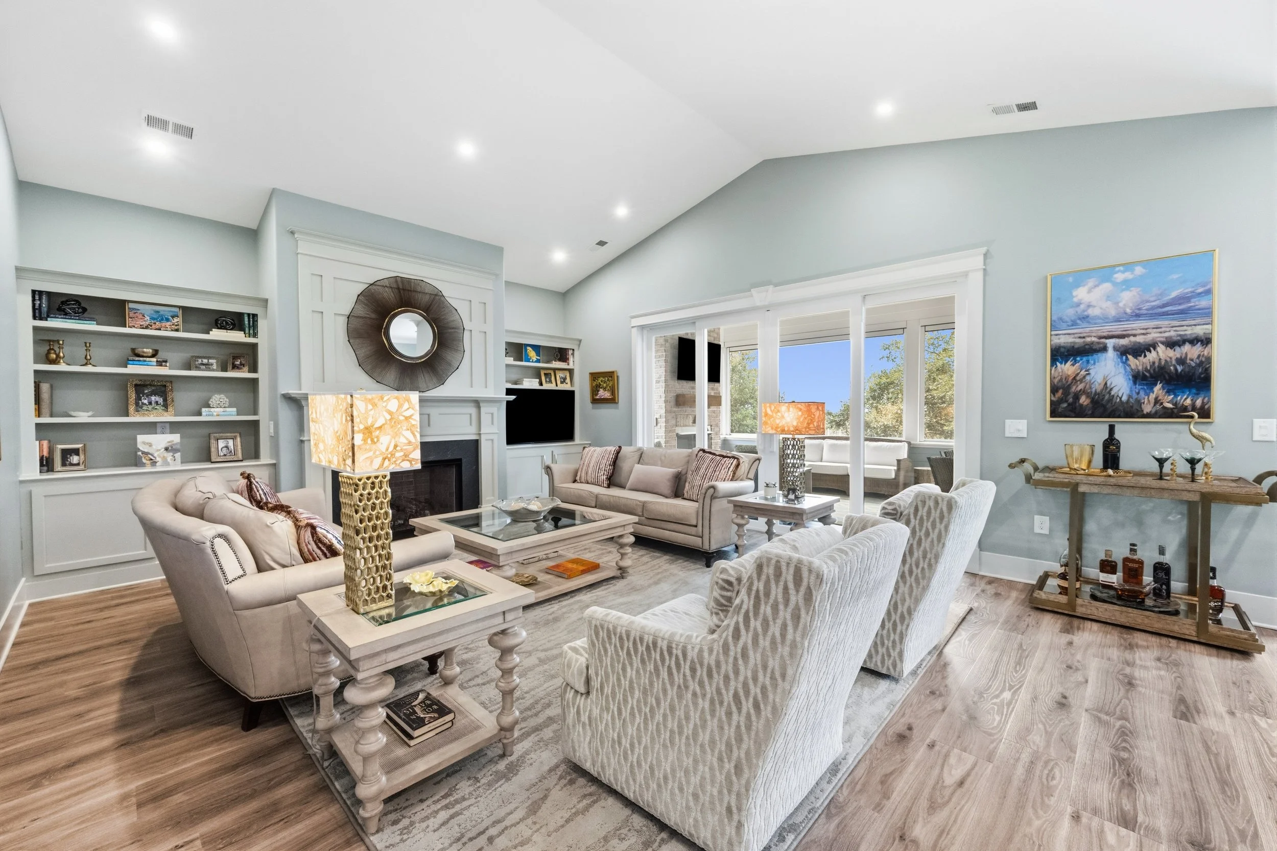 Living room with light blue walls, white crown molding, and wooden flooring. Features include a fireplace with a decorative mirror above it, built-in bookshelves, a beige armchair, a sofa, a coffee table, and a larger painting of a landscape. Large windows show trees outside, with sunlight coming in.